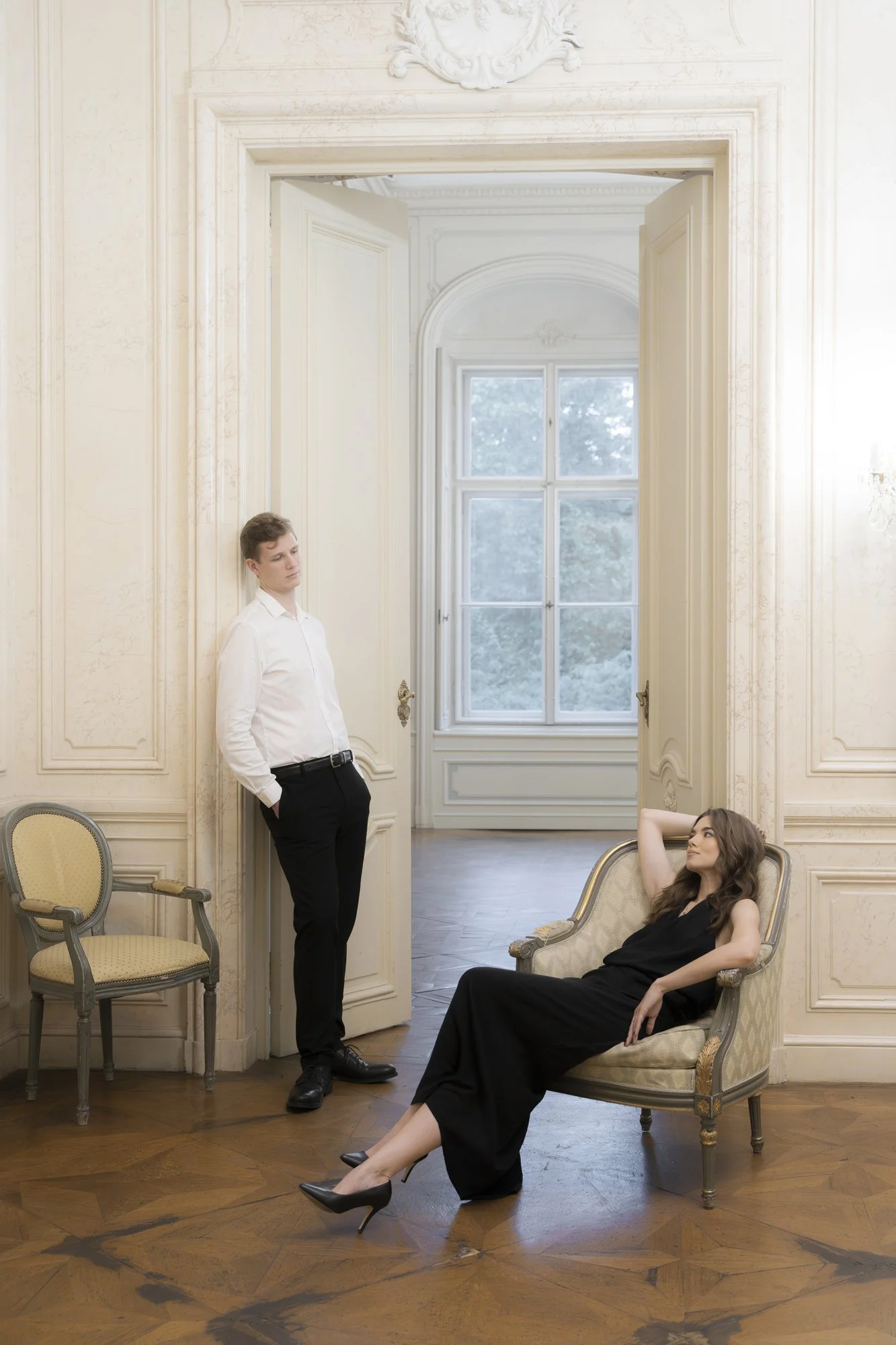 A woman in a black dress lounging on a vintage armchair with her legs crossed, and a man in a white shirt and black pants standing by the door in a luxurious room with ornate white walls and large windows.
