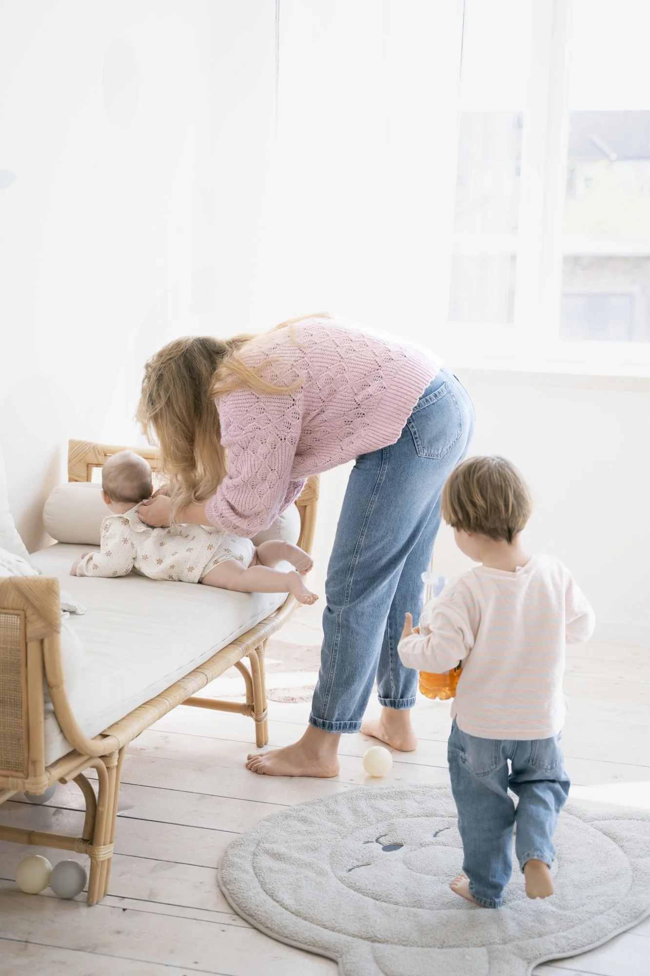A woman helping two young children in a bright, white room with wooden floors. One child is on a small bed, and the other is standing nearby holding a toy.