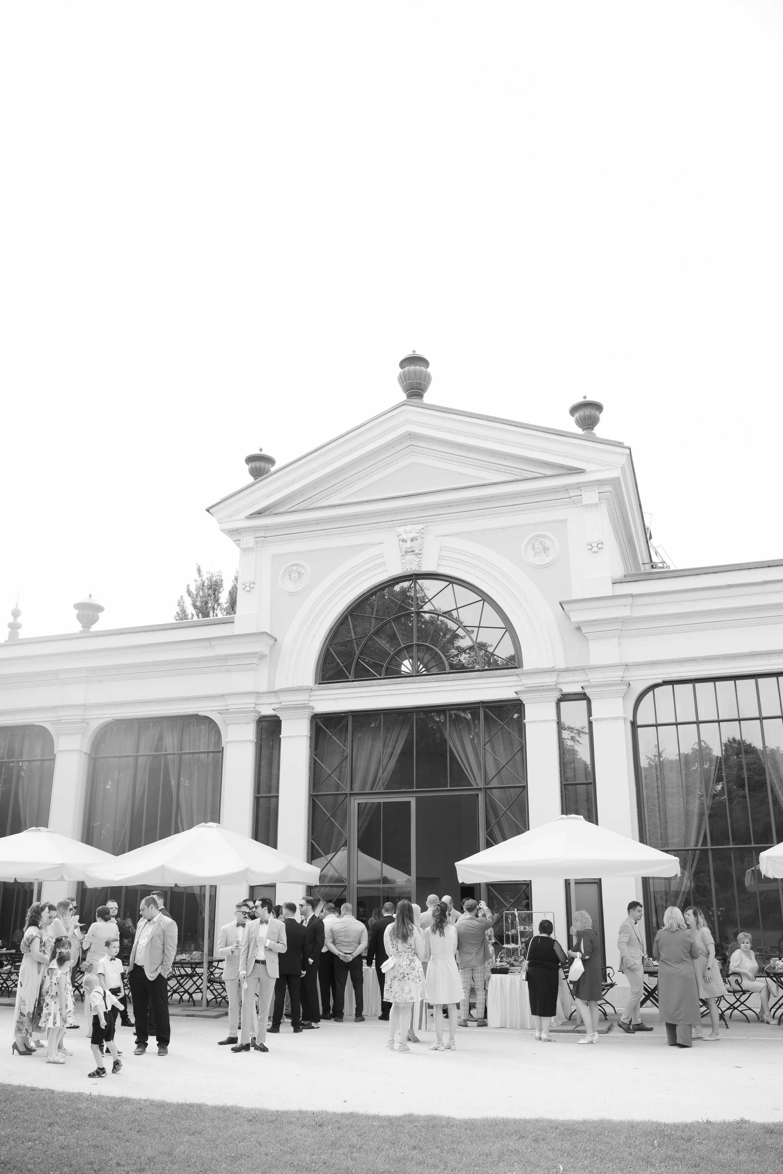 People gathering outside a large historic glass conservatory or building with umbrellas and tables, dressed in formal attire.