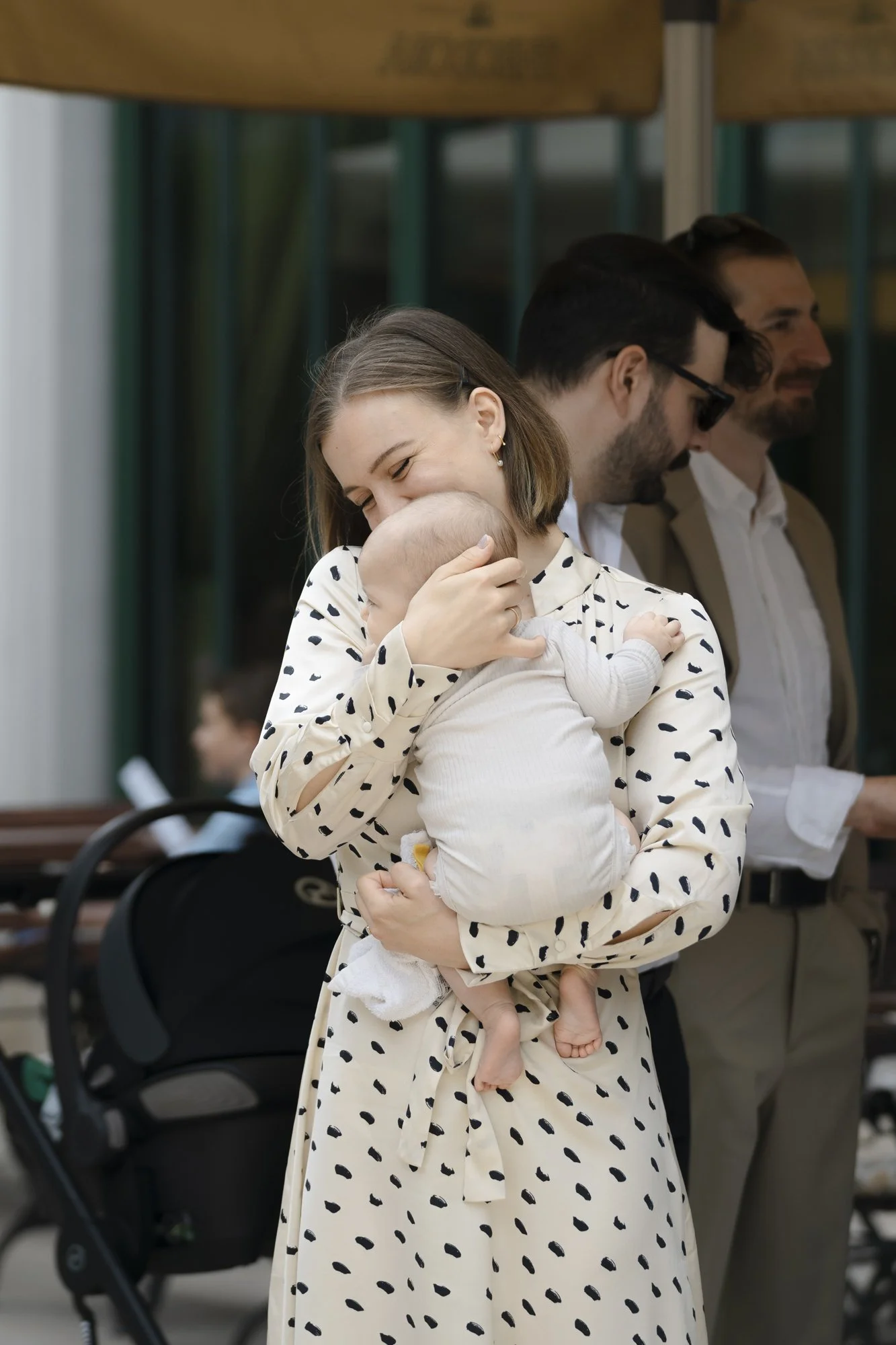 Woman holding a baby and smiling, standing near men in formal wear, at an outdoor event.