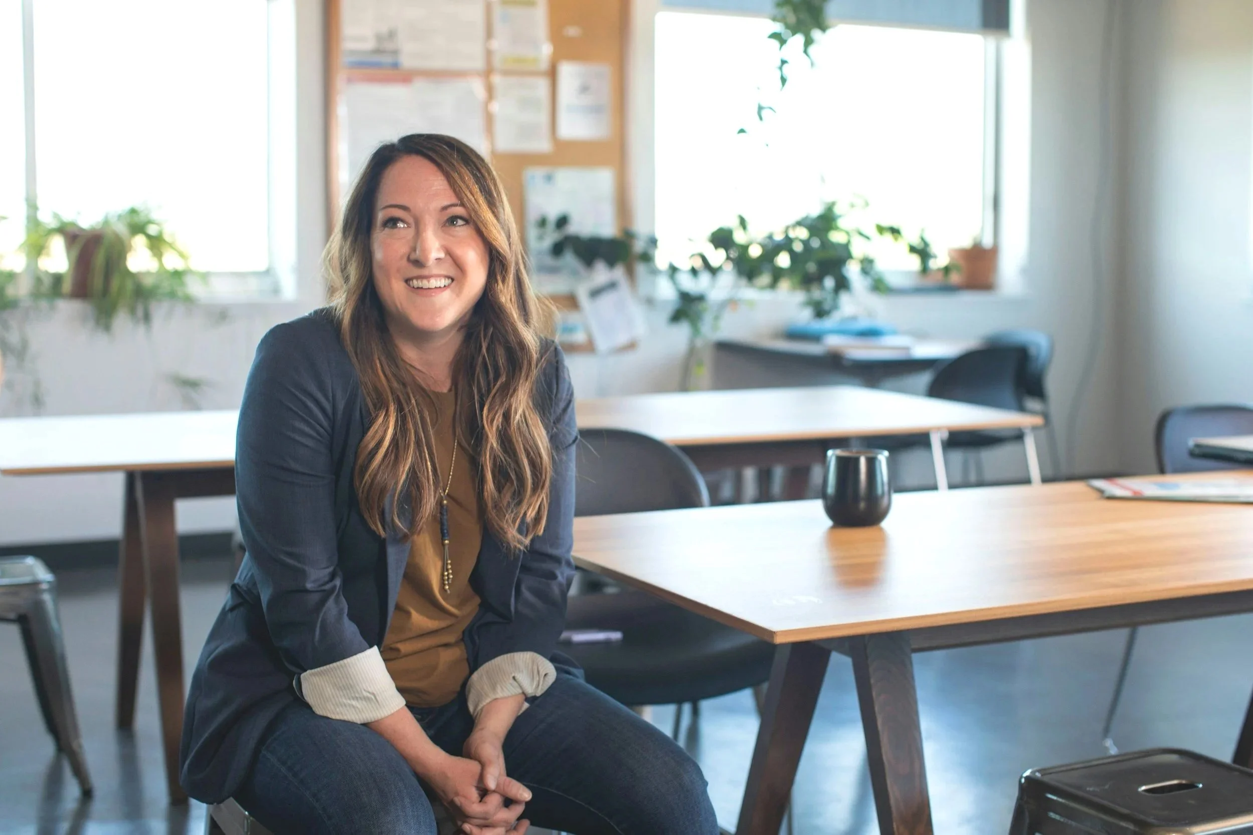 A woman with long brown hair smiling while sitting in a bright, casual office or classroom with windows, desks, and plants.