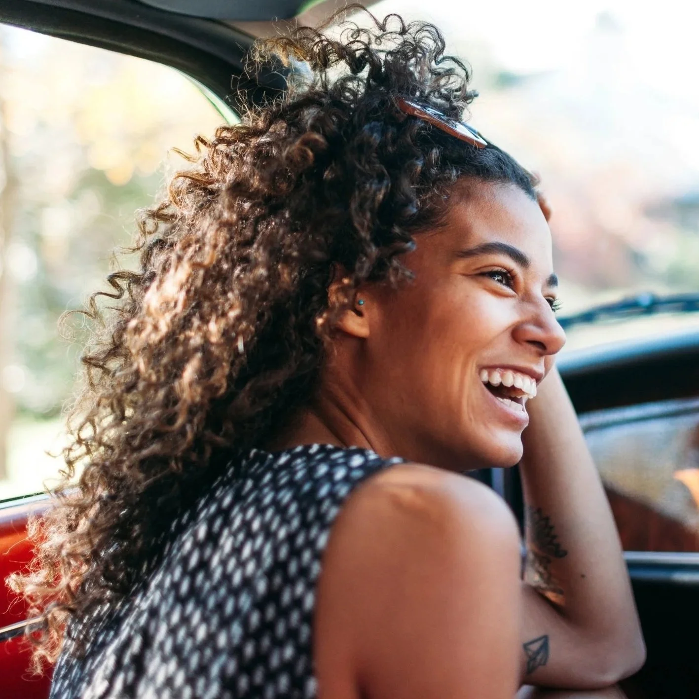 A young woman with curly dark hair smiling while sitting in a car, with sunlight in the background.