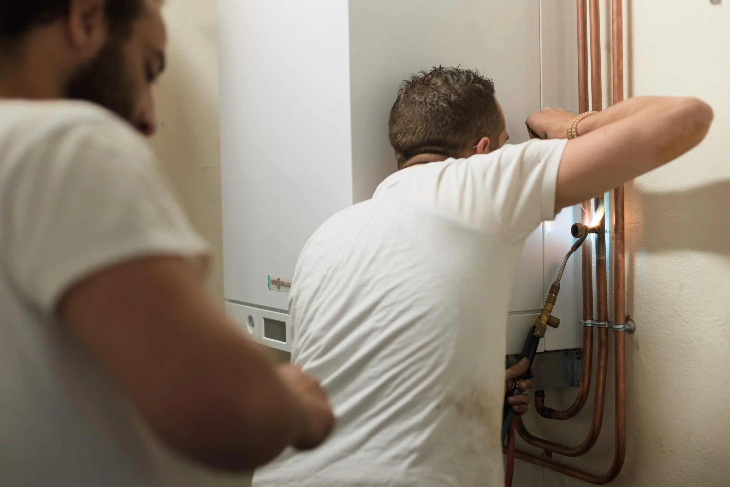 Deux hommes en blanc effectuent une réparation sur des tuyaux de plomberie copper dans une pièce intérieure.
