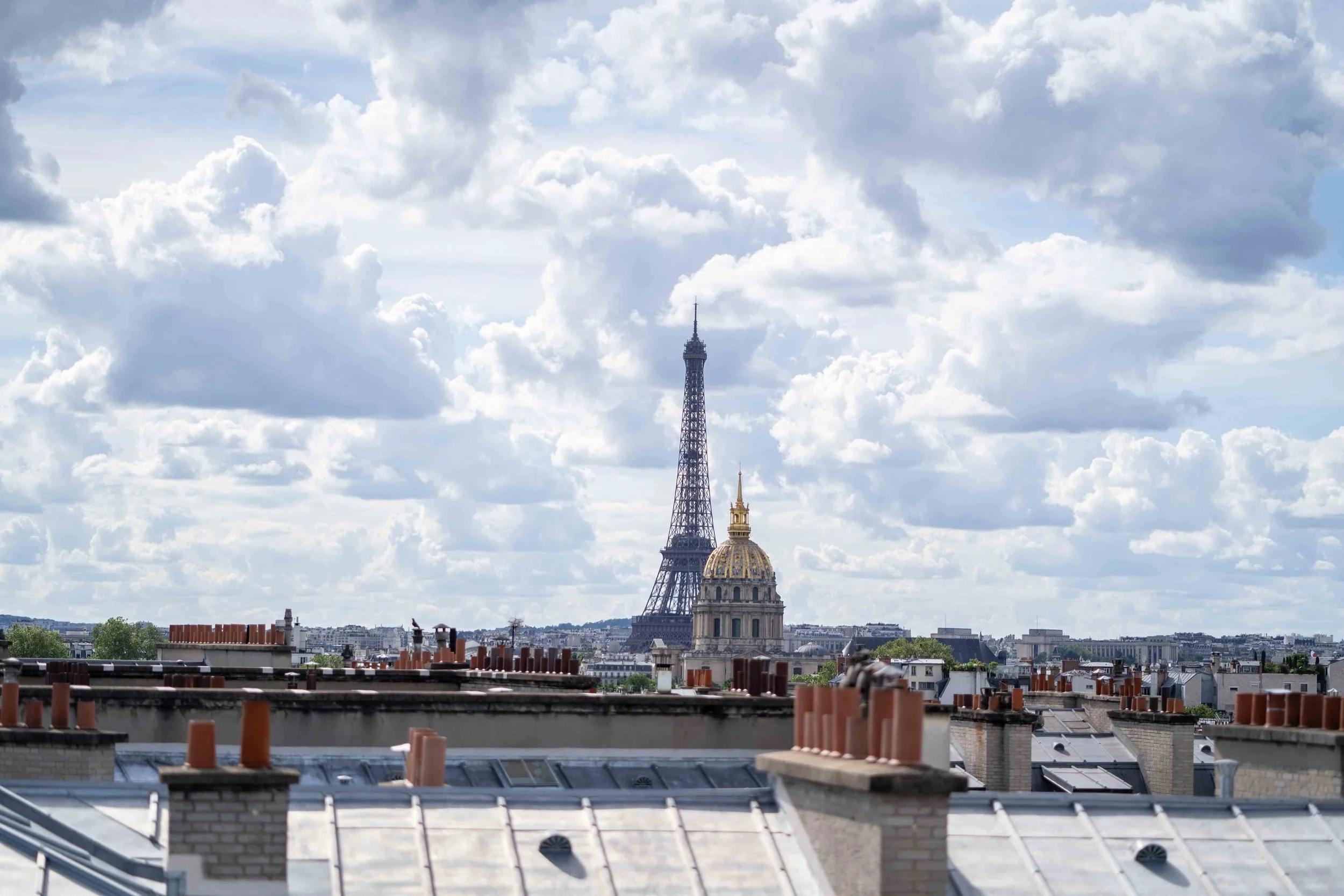 Vues de Paris avec la Tour Eiffel en arrière-plan, visible au centre de l'image, ainsi que plusieurs toits de bâtiments avec des cheminées en premiers plans.
