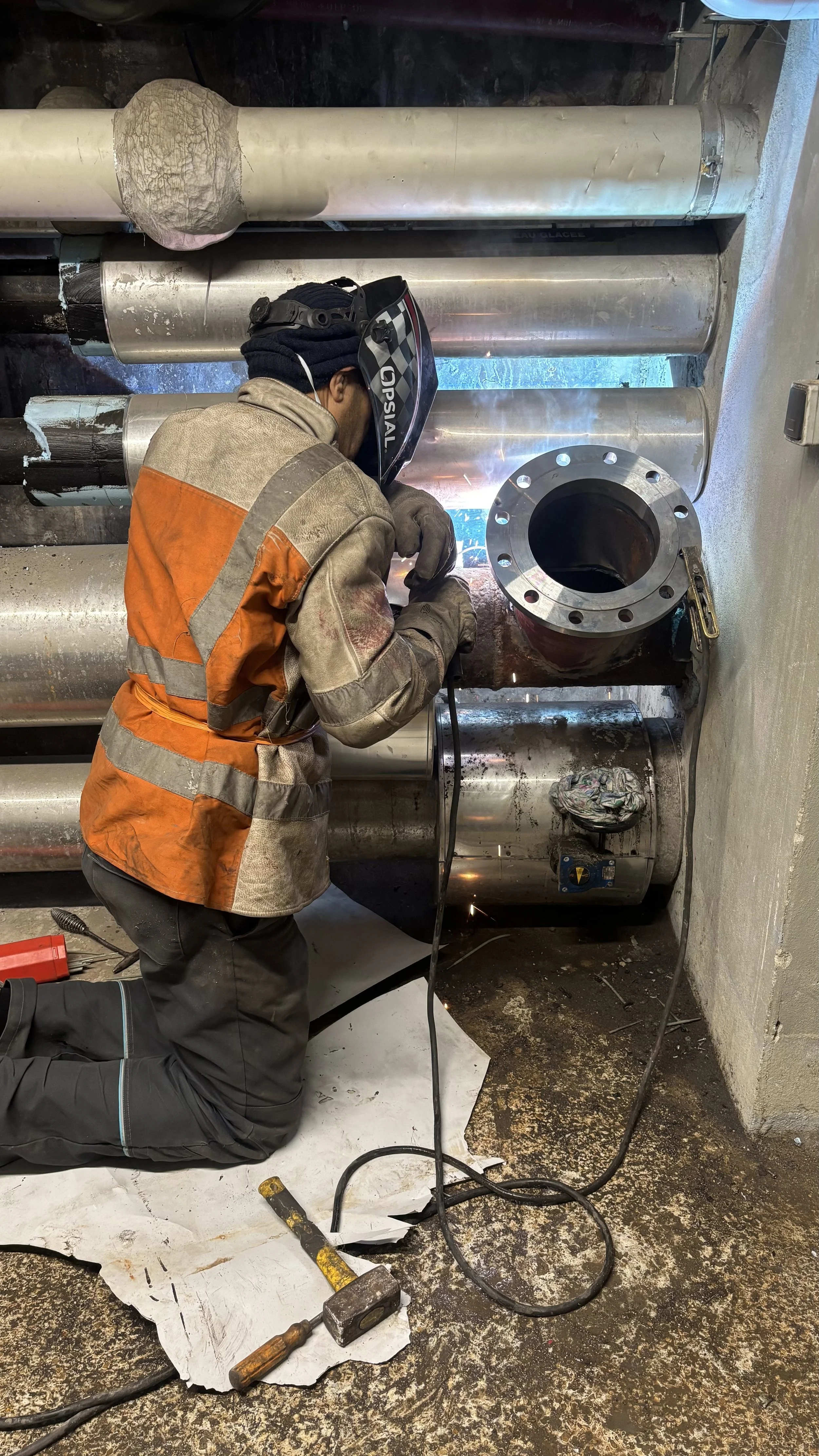 Un travailleur en équipement de protection worker, écrasant une pièce métallique à l'aide d'un équipement de soudage ou de coupe, dans un environnement industriel avec des tuyaux en acier.
