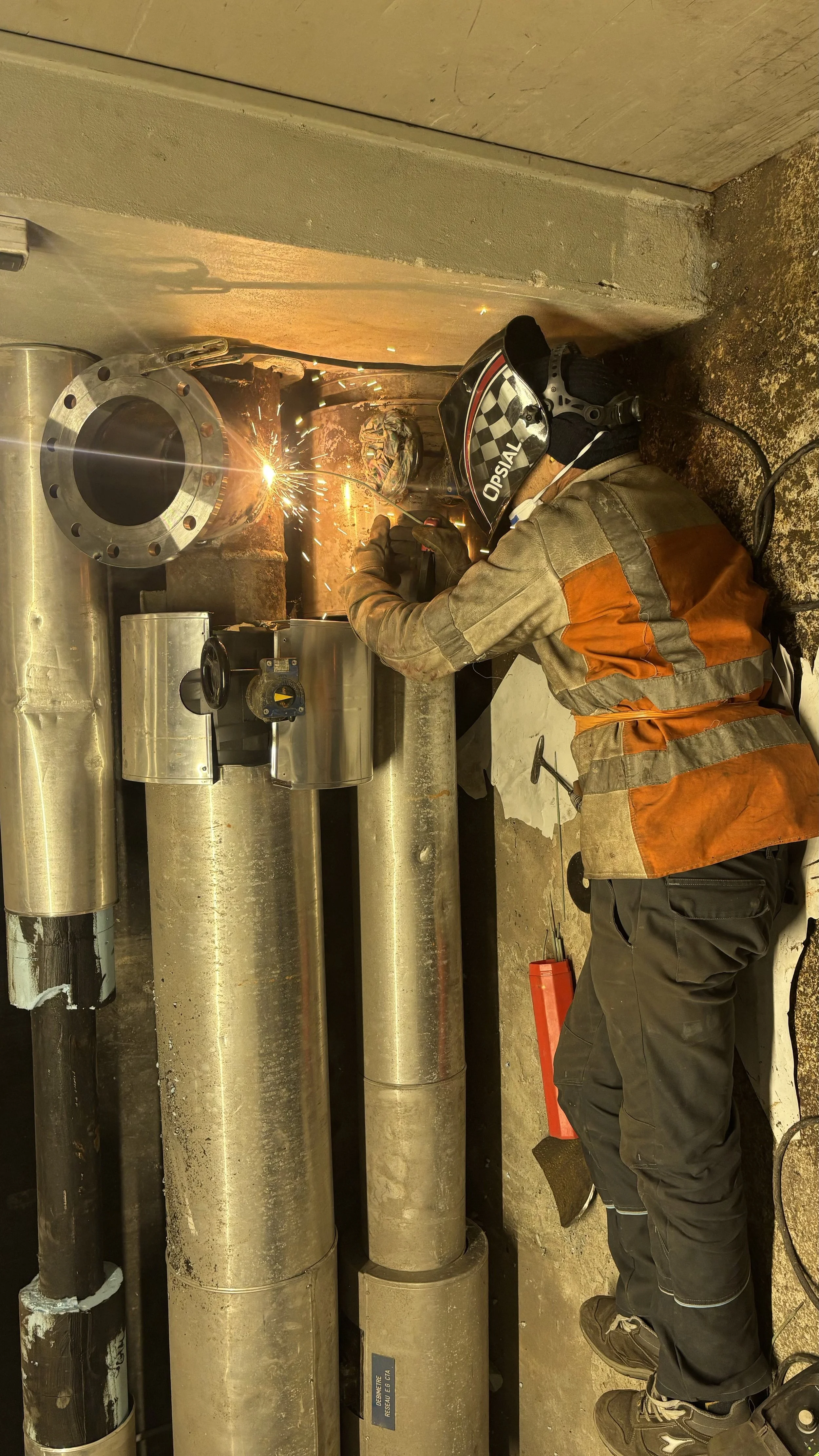 Un travailleur en équipement de sécurité, dont un casque de soudure, est en train de souder dans une installation industrielle ou de plomberie, avec des tubes métalliques et des équipements autour.