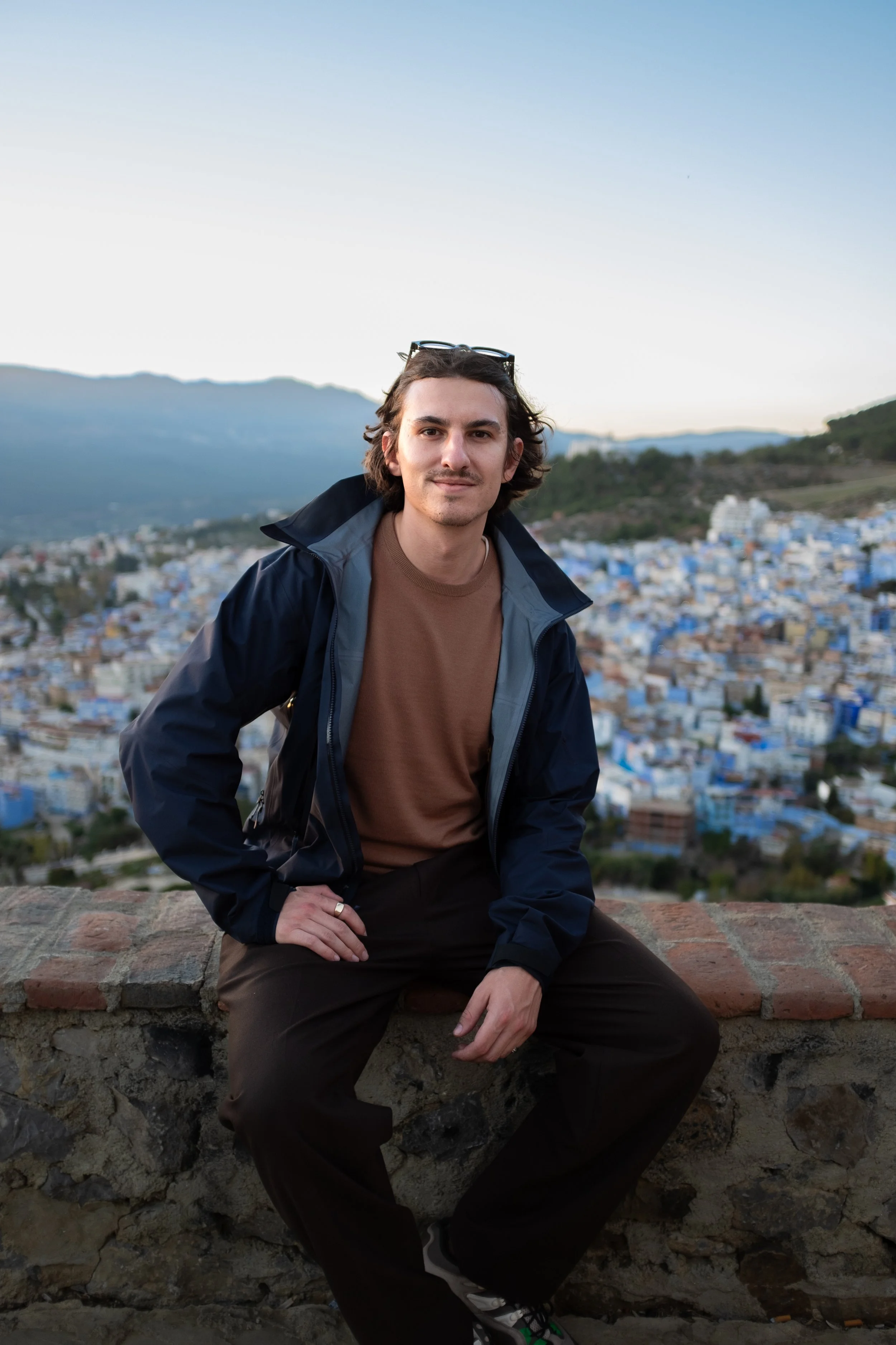 A young man with long dark hair and a slight mustache, wearing a brown shirt and a dark jacket, sitting on a stone wall with a cityscape behind him. The city features numerous blue buildings and a mountainous landscape.