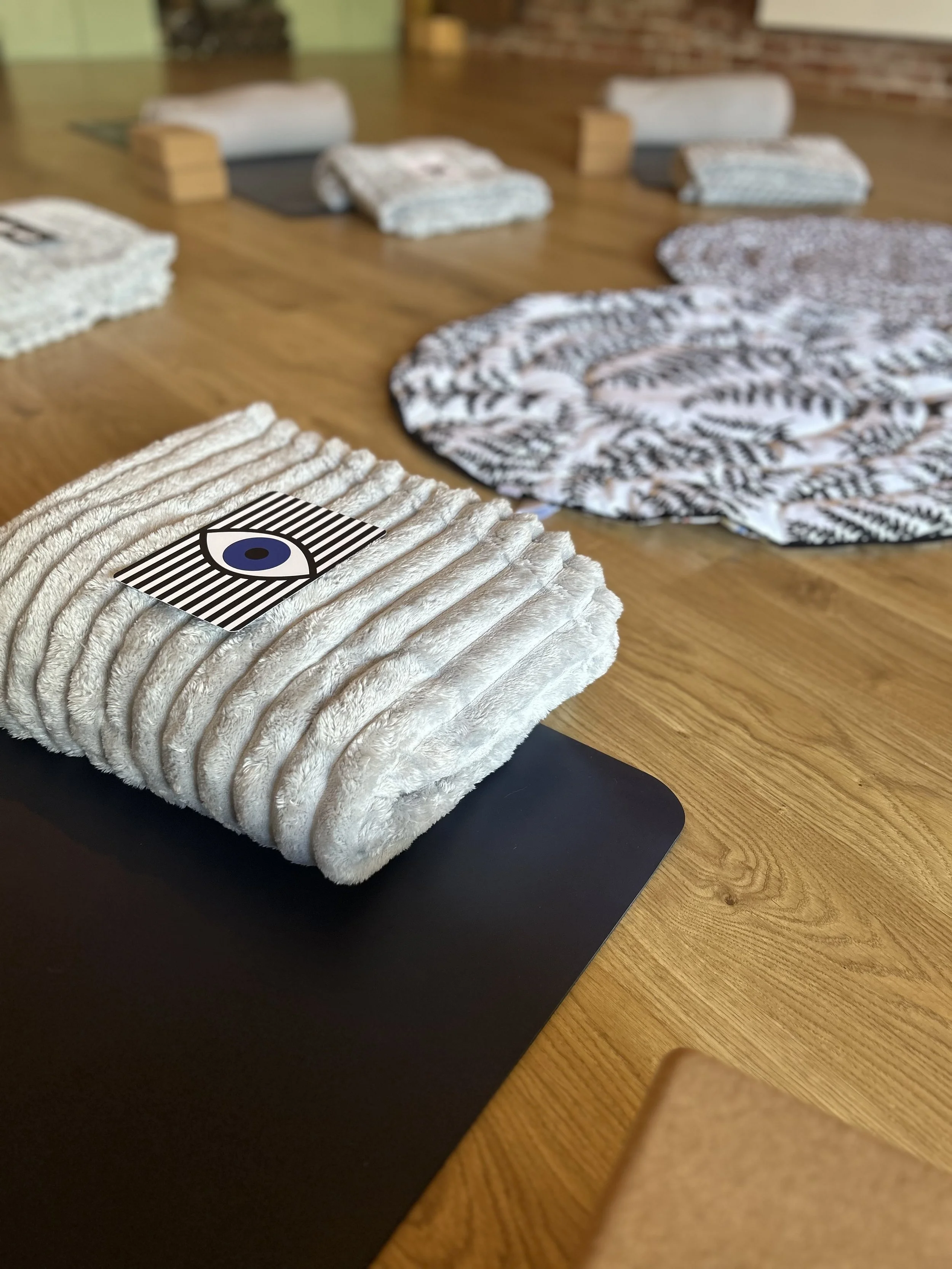 Close-up of a rolled, plush, beige towel with black and white barcode and eye design sticker on top for babies, on a black yogalina mat on a wooden floor, with folded blanket and a round patterned baby kick mat in the background.