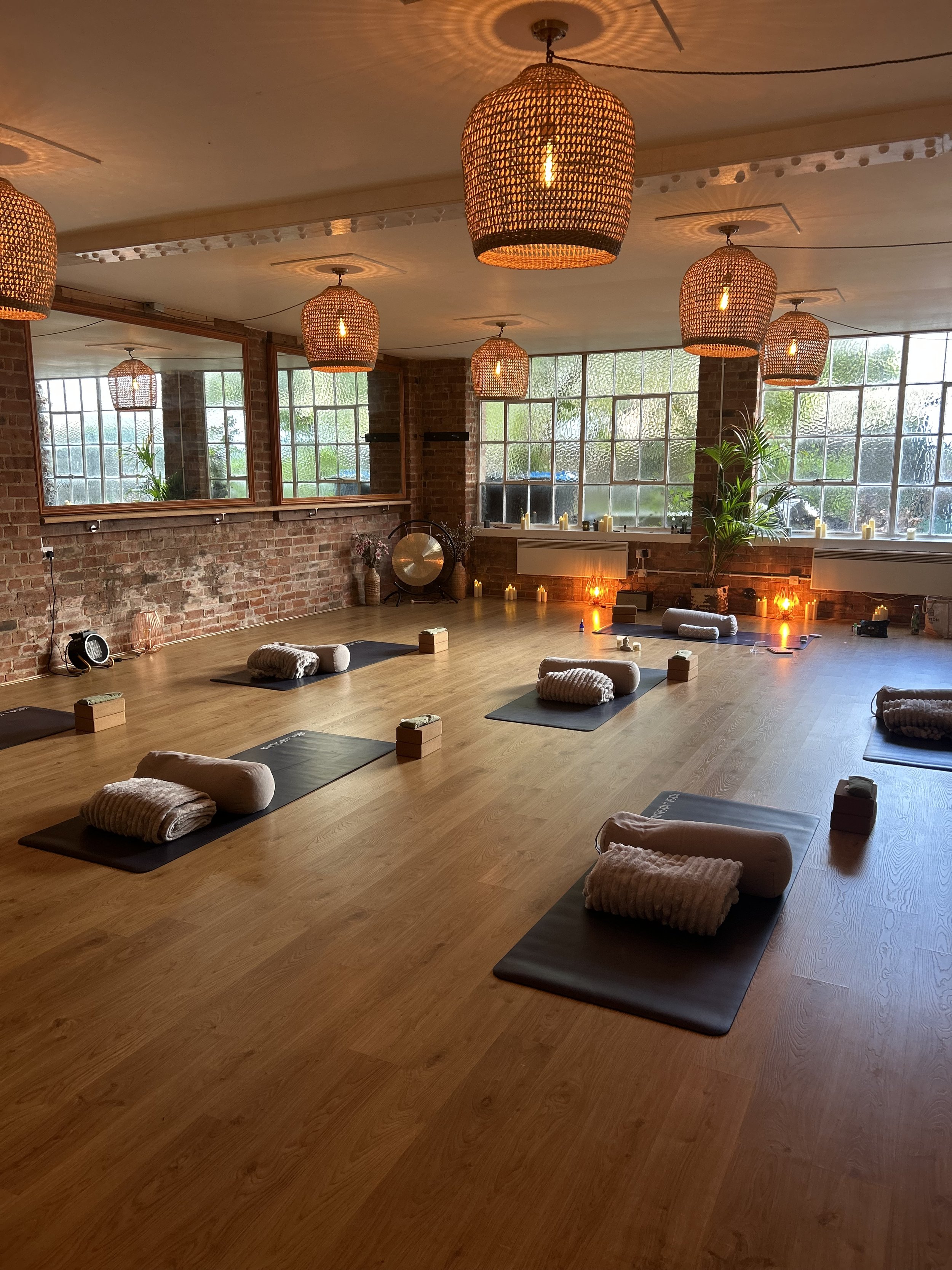 Yoga studio in Witney with wooden floors, large windows, and hanging woven pendant lights. Yoga mats with bolster pillows and blocks are spaced evenly on the floor. Candles and decorative items are near the windows to create a warm cosy environment.