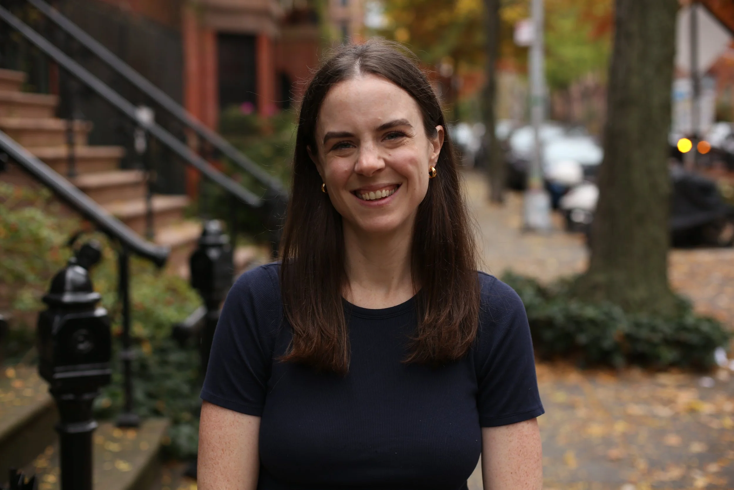 A woman with long brown hair and freckles smiling in an outdoor urban setting with trees, houses, and parked cars in the background during fall.