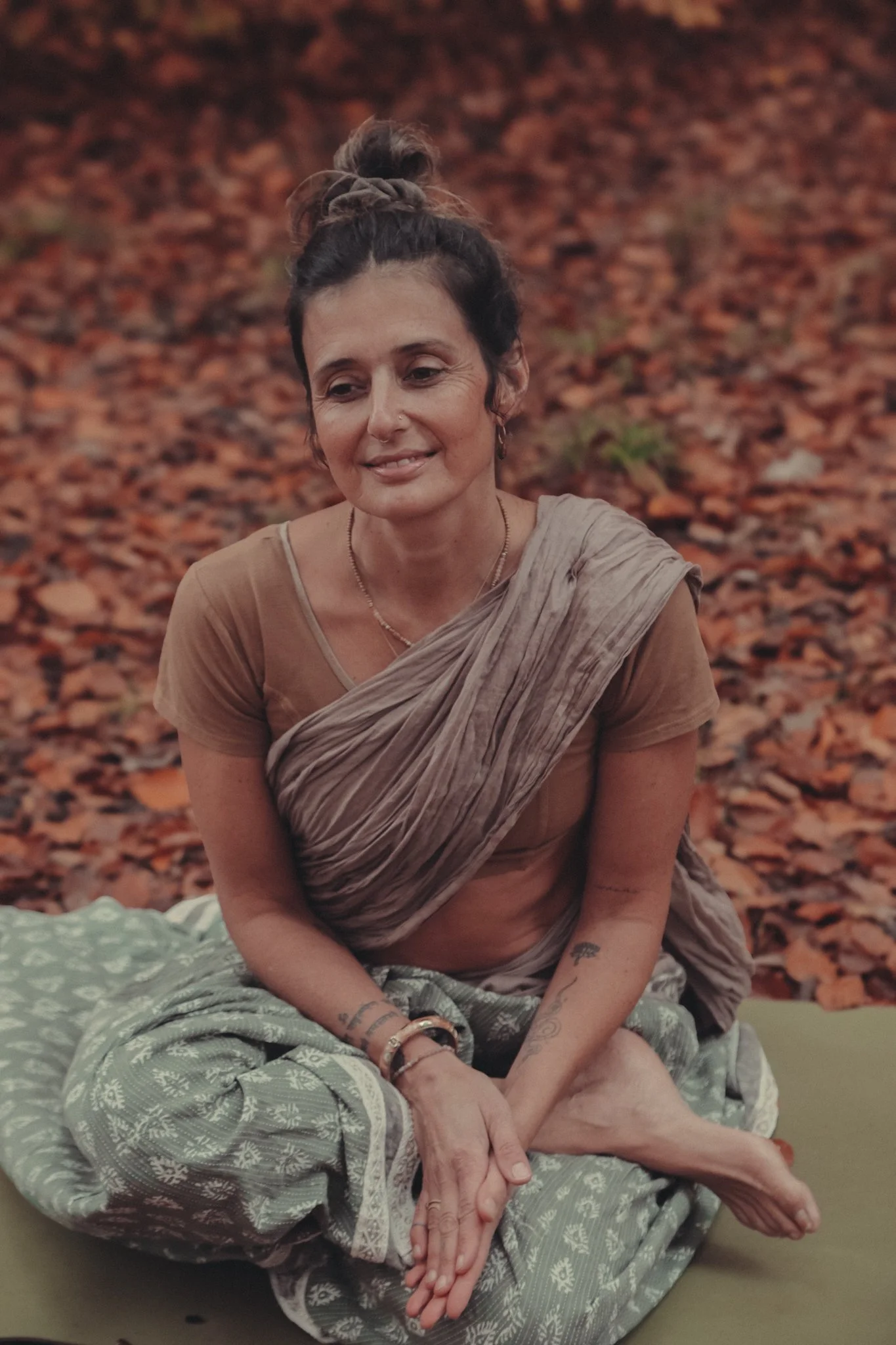 Eine Frau mit dunklen, gewellten Haaren in einem Haarknoten ist entspannt und lächelt leicht, während sie in einem Raum mit Herbstlaub im Hintergrund sitzt.