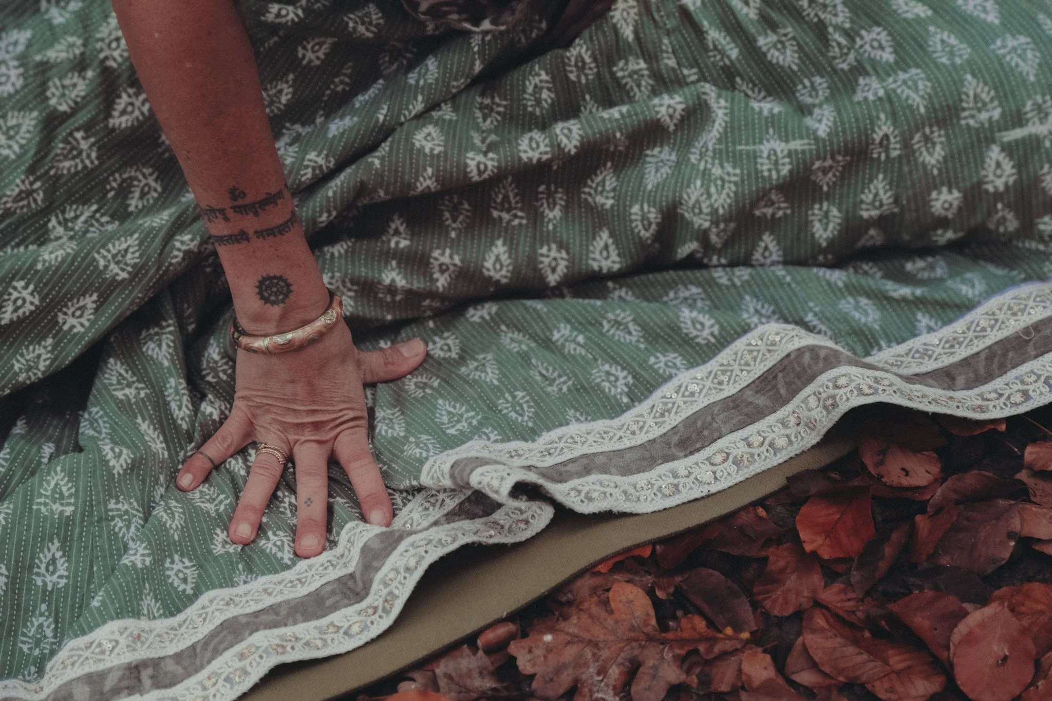 Hand und Fuß einer Person mit Tattoo auf dem Arm, die auf bunten Decken liegen, umgeben von Herbstblättern.