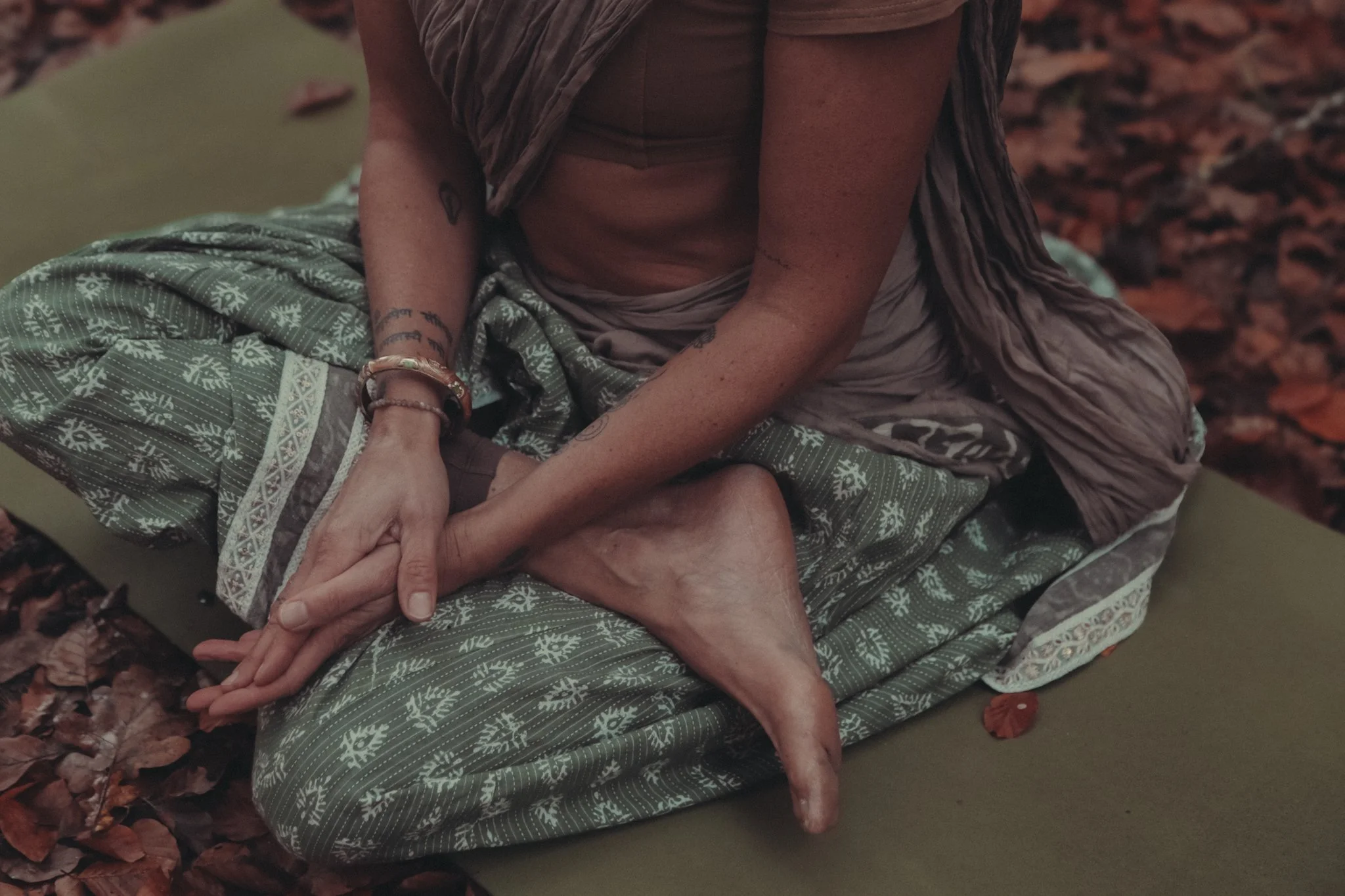 Eine Person sitzt auf dem Boden, umgeben von Herbstblättern, mit sichtbaren Tätowierungen und bunten Armbändern, trägt bequeme Kleidung mit Pflanzenmuster.