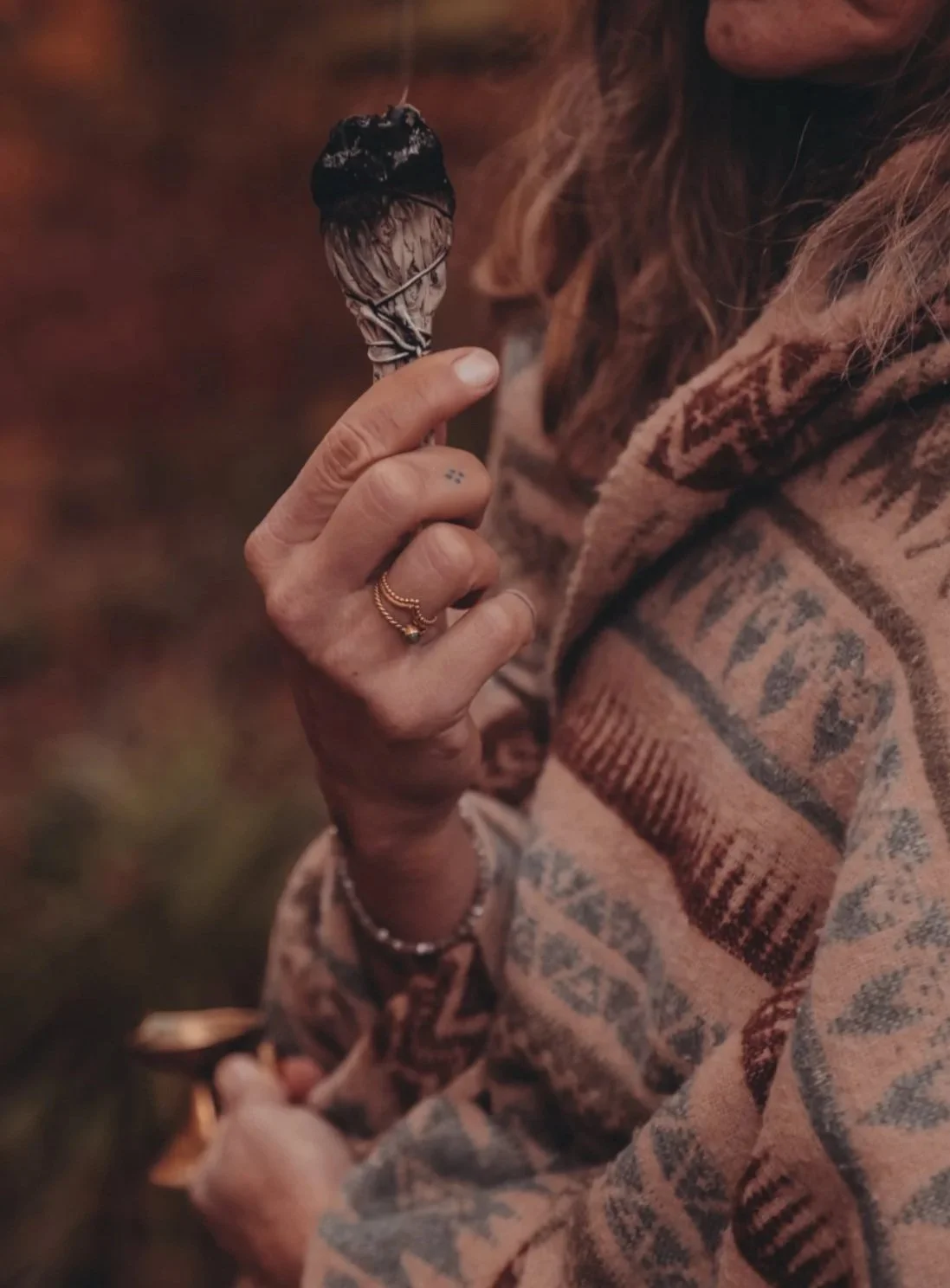 Person hält eine halb verbrannte Zigarette in der Hand, trägt goldene Ringe und Armbänder, trägt ein kariertes, buntes Hemd, mit langen Haaren.