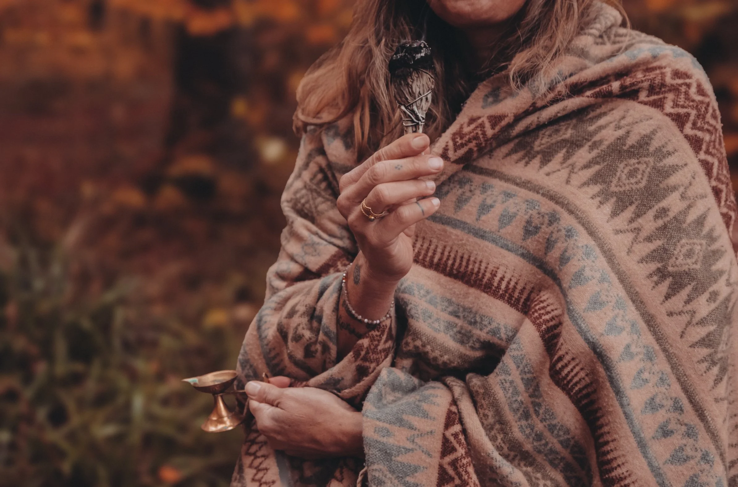 Eine Person trägt ein großes, gemustertes Tuch und hält eine kleine Kerze in der Hand. Die Person hat lange, gewellte Haare. Der Hintergrund ist unscharf mit Herbstlaub.