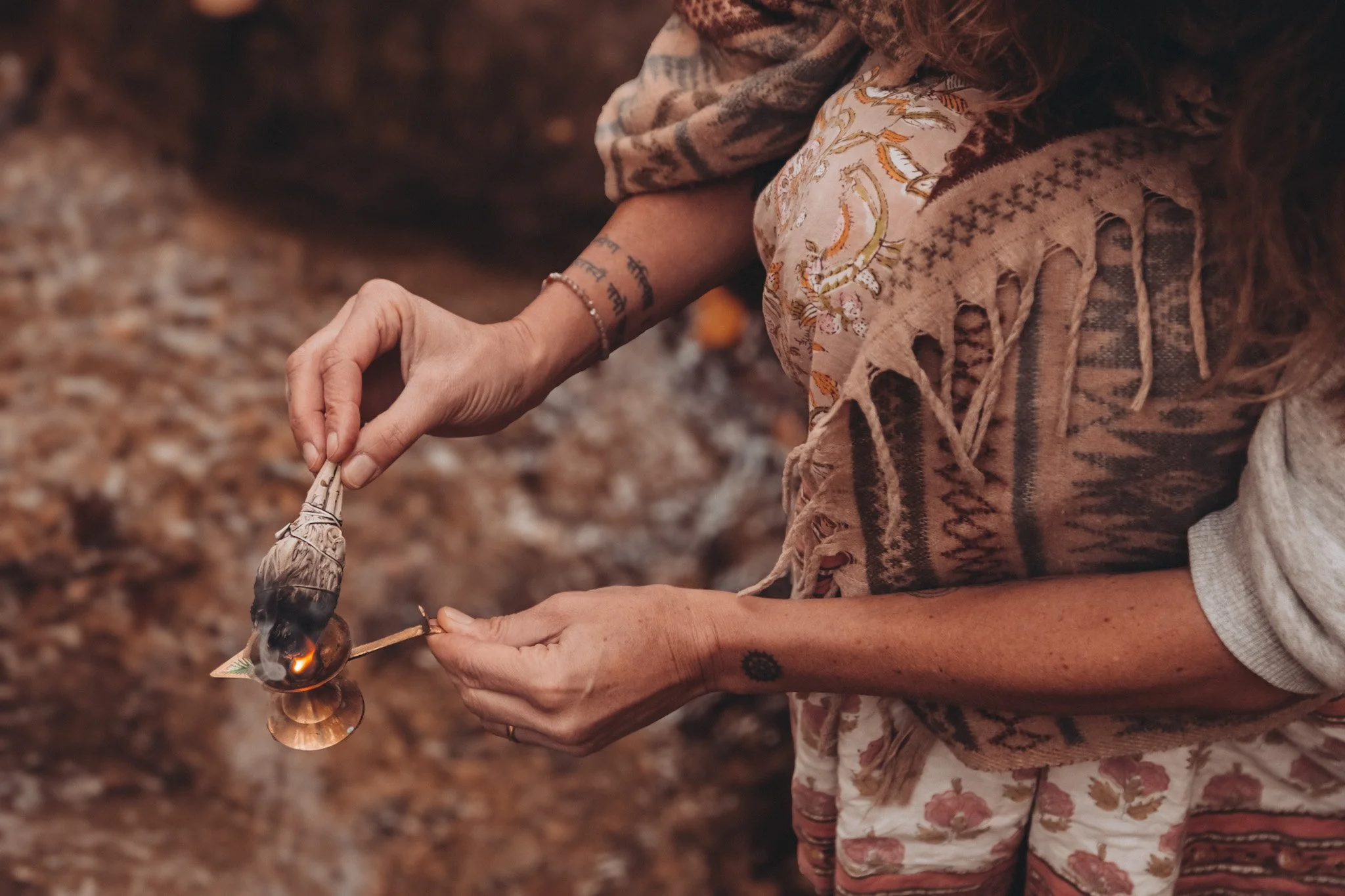Nahaufnahme einer Person, die eine Kerze anzündet. Die Person trägt einen bunten Schal und eine Bluse mit Blumendruck, hat Tattoos am Unterarm und trägt Schmuck. Die Umgebung ist unscharf und erinnert an einen Herbstwald.