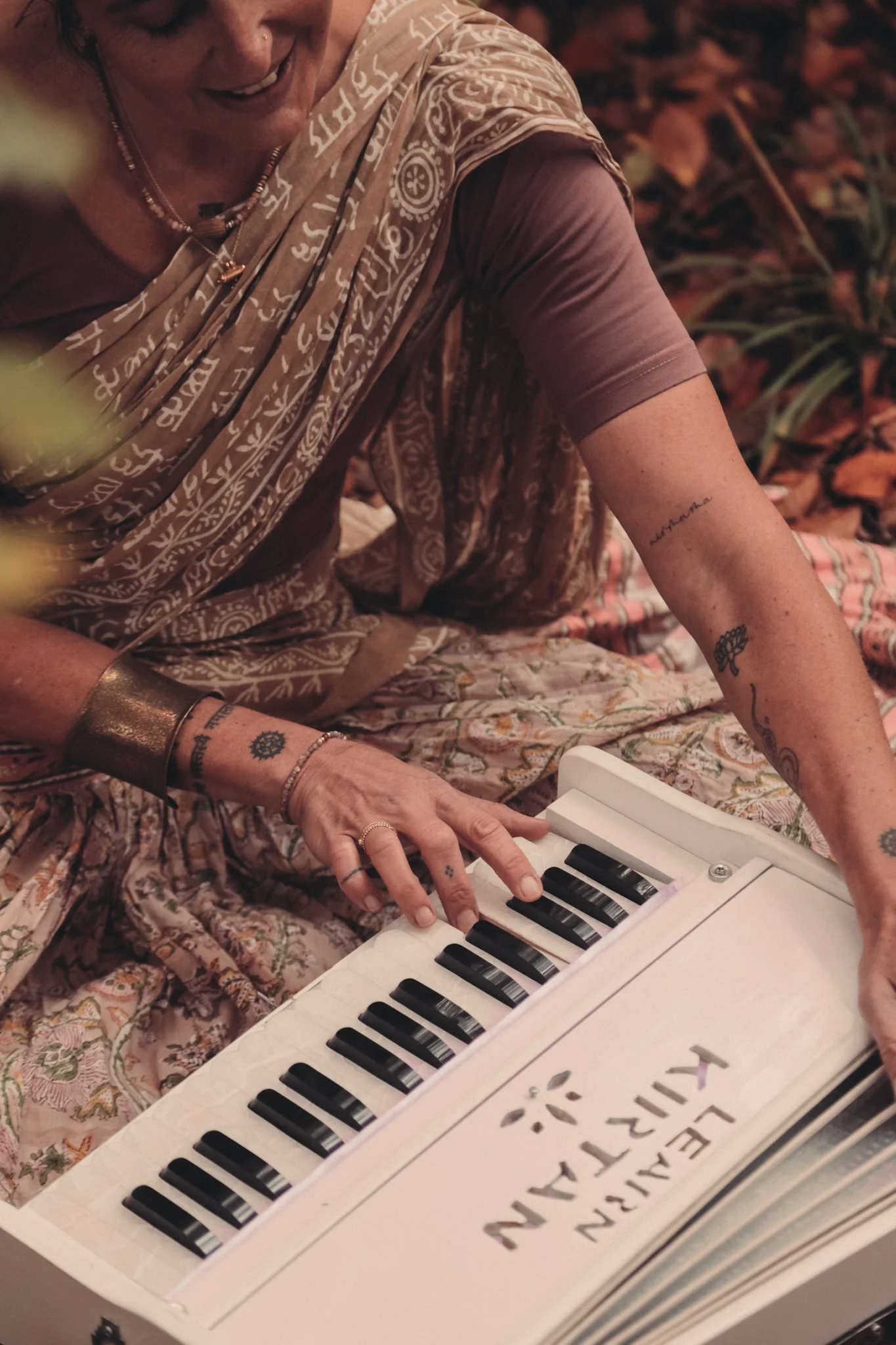 Eine Frau spielt auf einem kleinen weißen Spielzeugklavier mit schwarzen und weißen Tasten. Sie trägt ein blumenmusters Tuch und Schmuck. Im Vordergrund ist die Seite des Klaviers mit aufgedrucktem Text sichtbar.