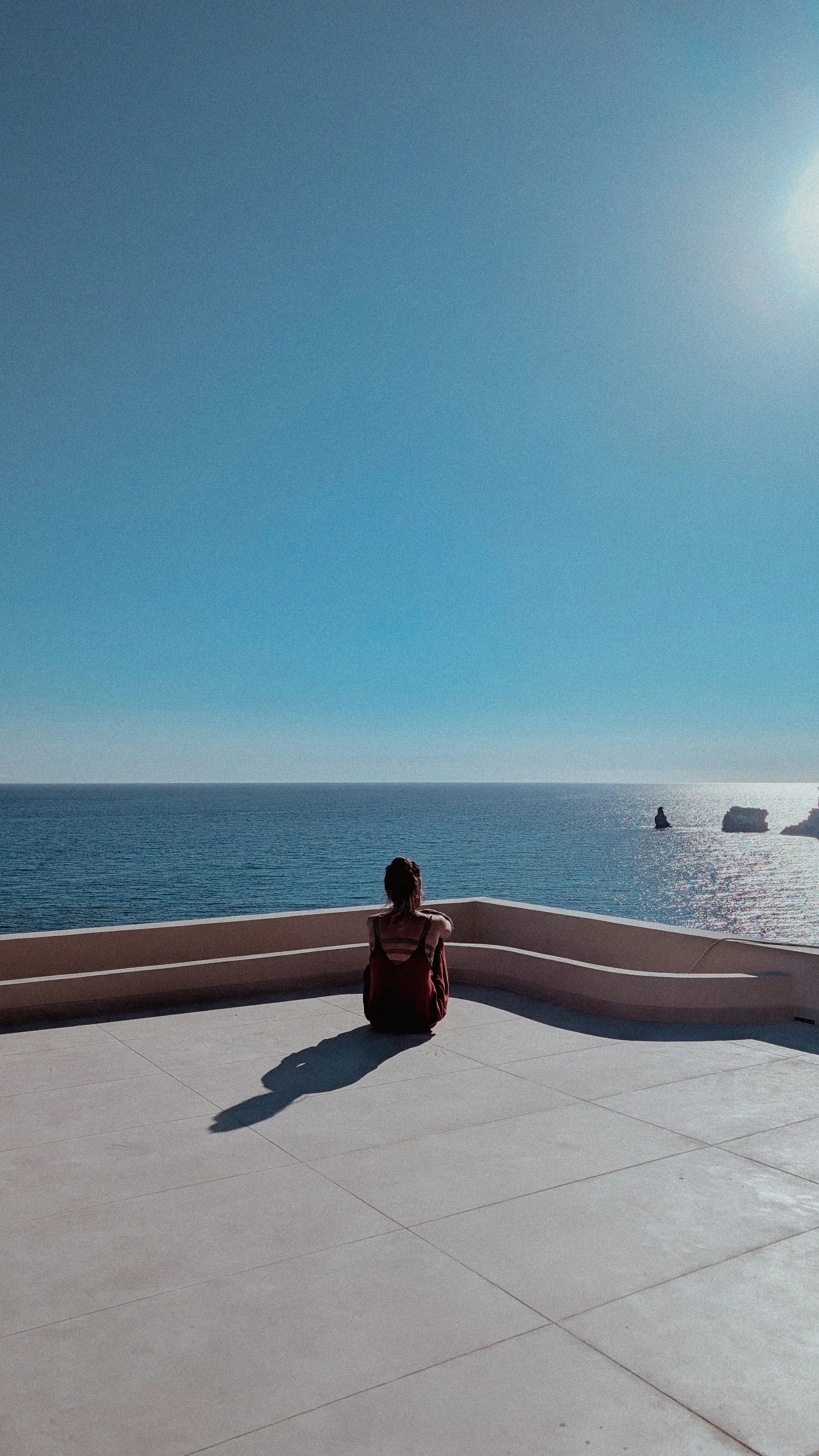 Eine Frau sitzt auf einer Terrasse, blickt auf das Meer und den Himmel während des Sonnenuntergangs, mit Blick auf Felsen im Wasser.