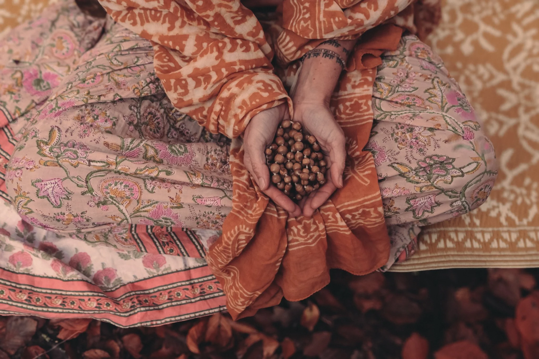 Eine Person in bunten, mehrlagigen, traditionell gemusterten Kleidern hält eine Hand voll Haselnüsse. Die Person trägt ein orangefarbenes Oberteil mit weißen Mustern und einen Rock mit floralem Muster. Der Hintergrund besteht aus mehreren Schichten einer ähnlichen Kleidung mit floralem Design.