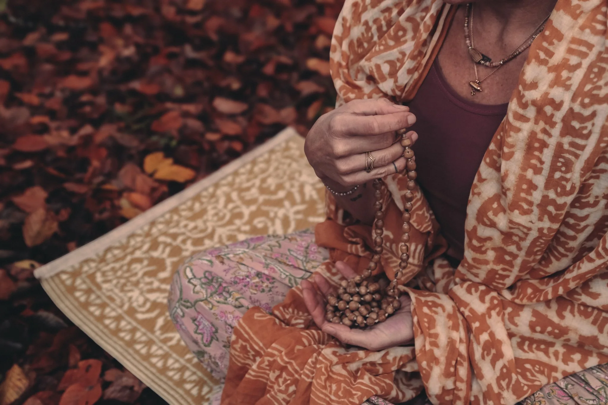 Nahaufnahme einer älteren Frau, die traditionelle indische Kleidung trägt, Schmuck hält und auf einem geblümten Kissen sitzt, umgeben von gefallenen Blättern.