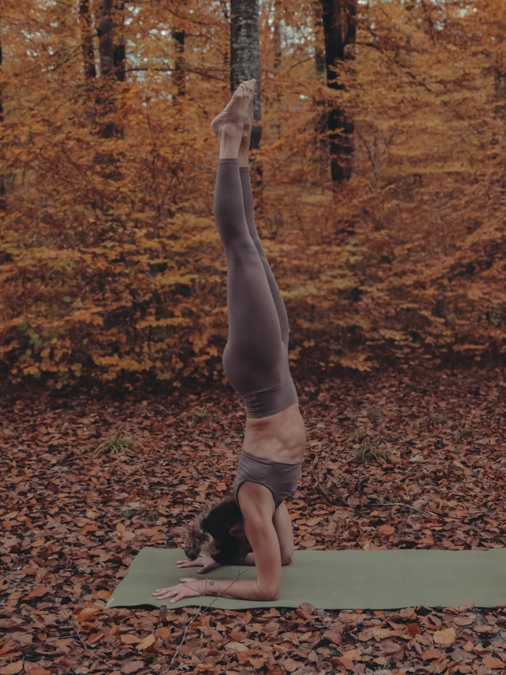 Frau macht einen Handstand auf einer Yogamatte im herbstlichen Wald mit orangefarbenen Blättern.
