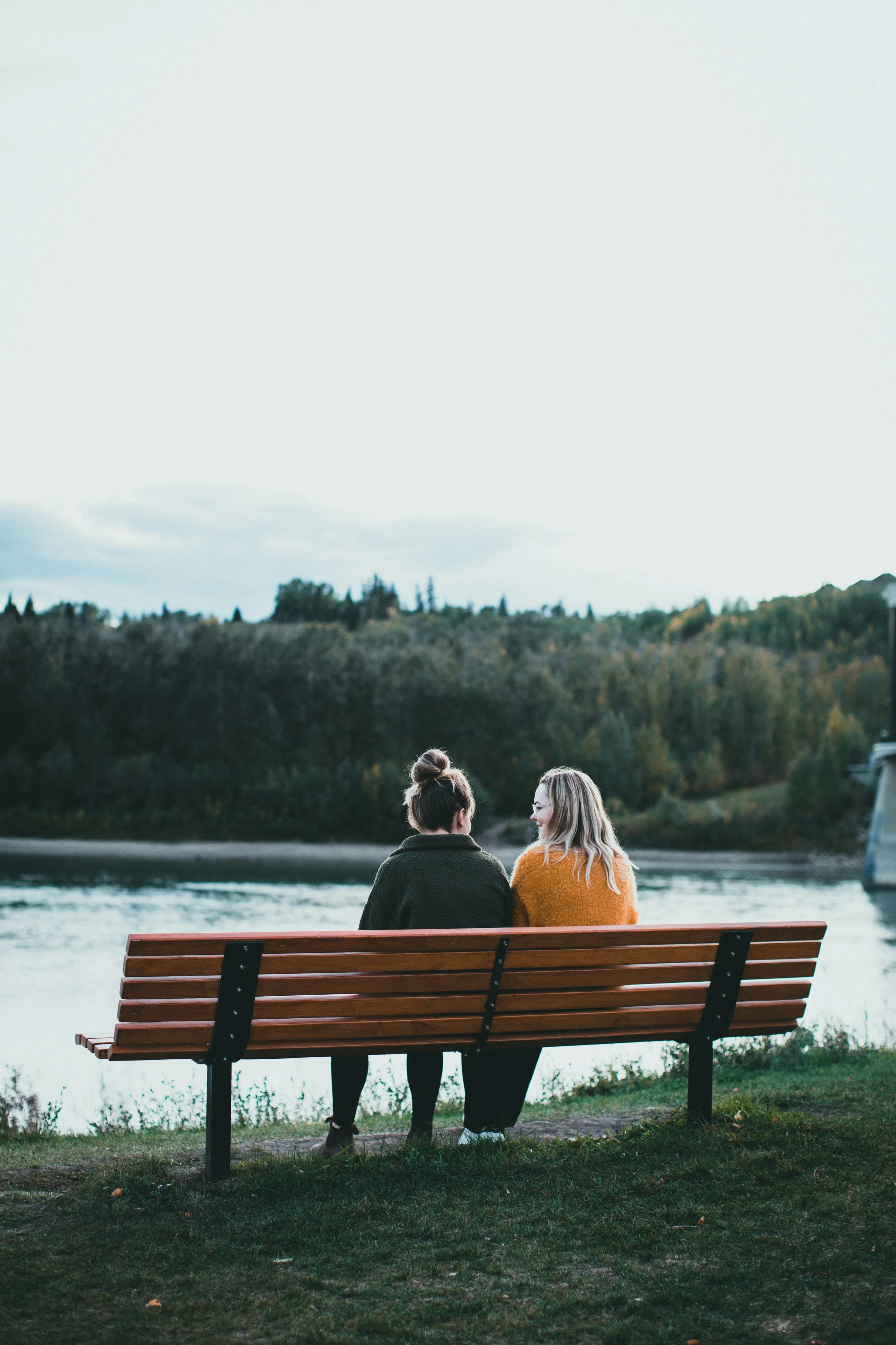 Two women sitting on a park bench by a river, facing away, engaging in a conversation, with trees and hillside in the background.