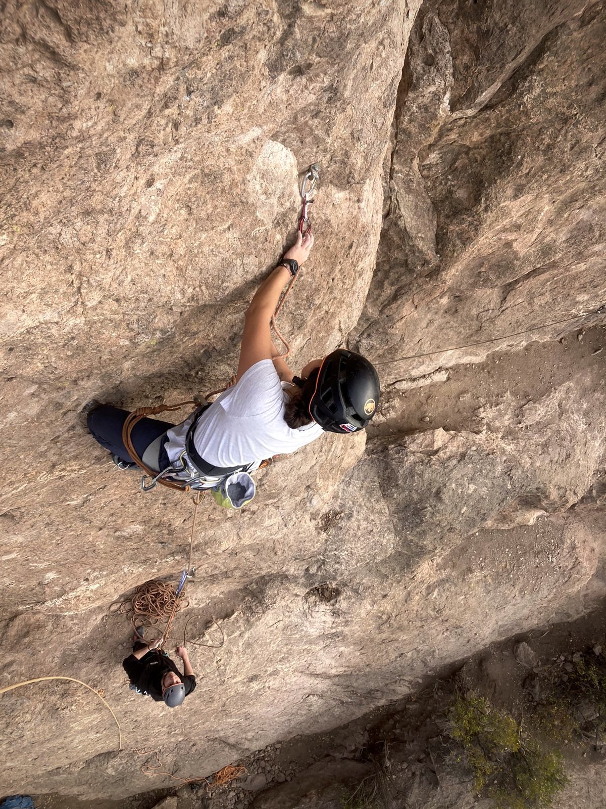 Person rock climbing on a vertical mountain wall, wearing a helmet and climbing gear, with a smaller person below, also wearing a helmet, secured by ropes.