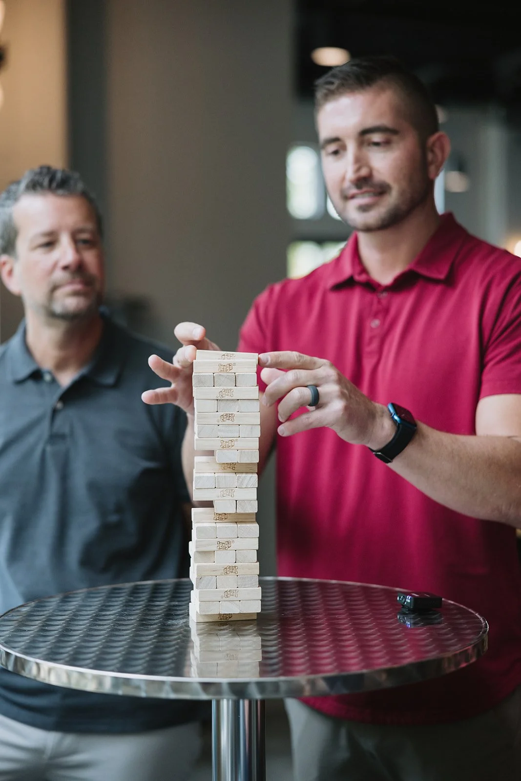 Two men playing Jenga, one in a red shirt balancing a wooden block on a tall Jenga tower, the other man watching.
