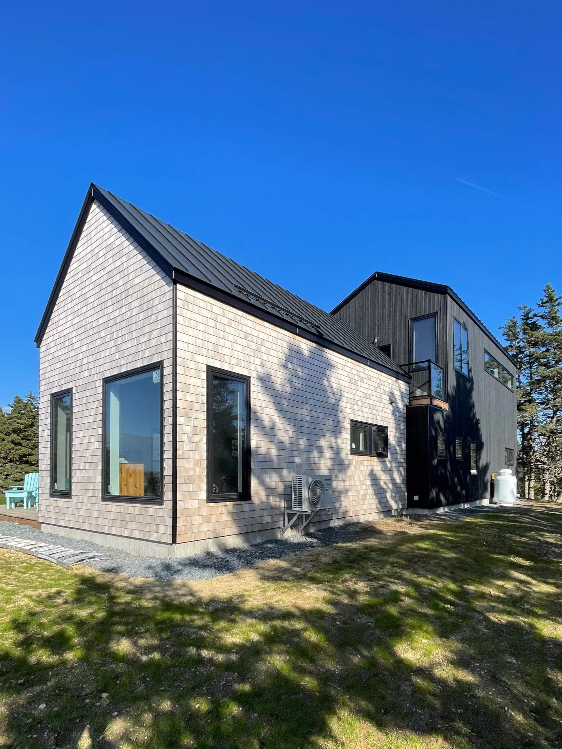 Modern house with wooden and black siding, large windows, located in a grassy yard under a clear blue sky with shadows of trees.