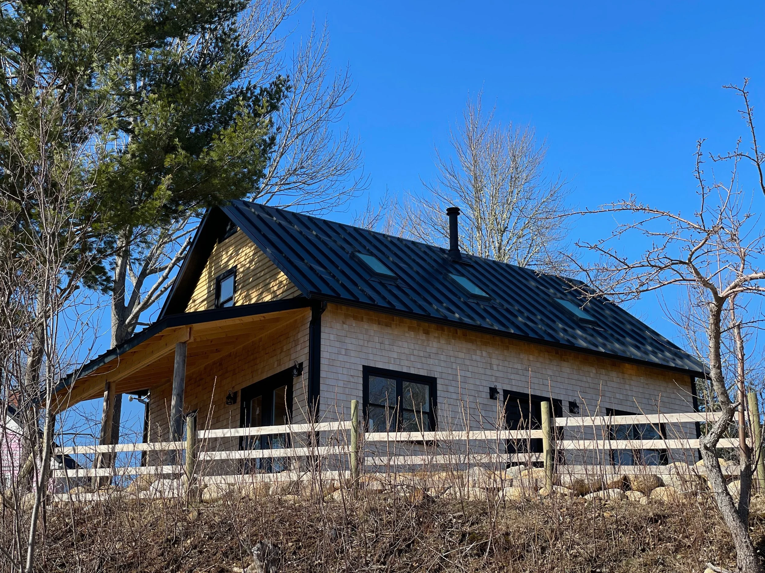 A two-story house with beige siding, black roof with skylights, black window frames, and a small front porch. The house is surrounded by leafless trees and a wooden fence, under a bright blue sky.