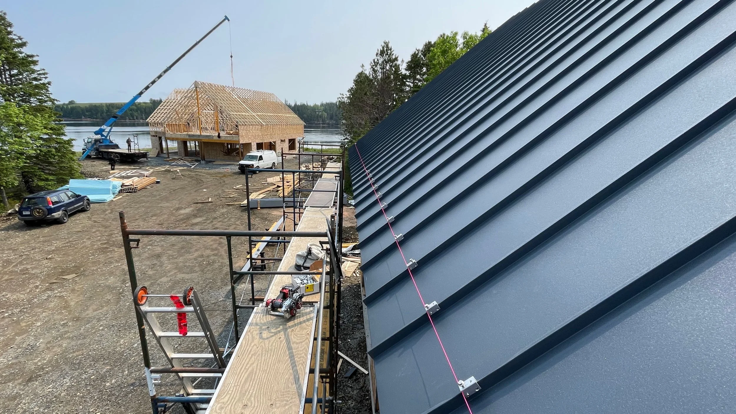 Construction site near a lake showing a partially built house with a crane, parked vehicles, and a new metal roof being installed on a building.