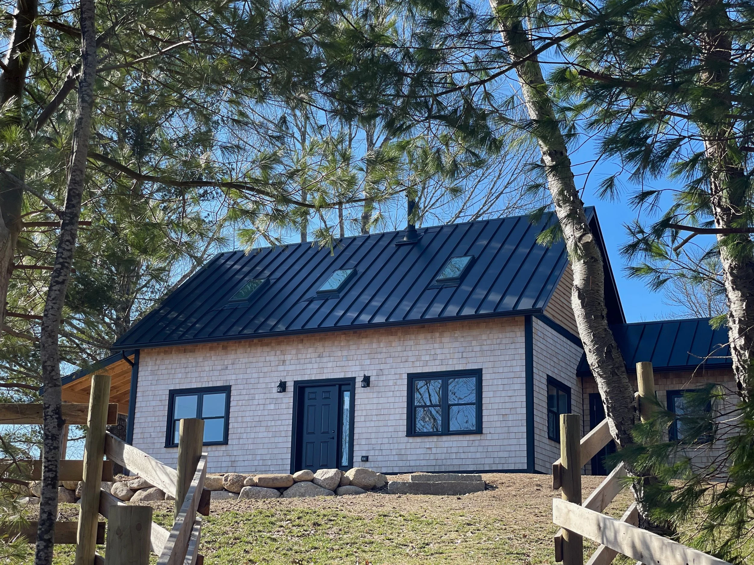 A new house with a dark metal roof, beige shingled exterior walls, and black window frames, situated among trees with a pathway leading up to it.