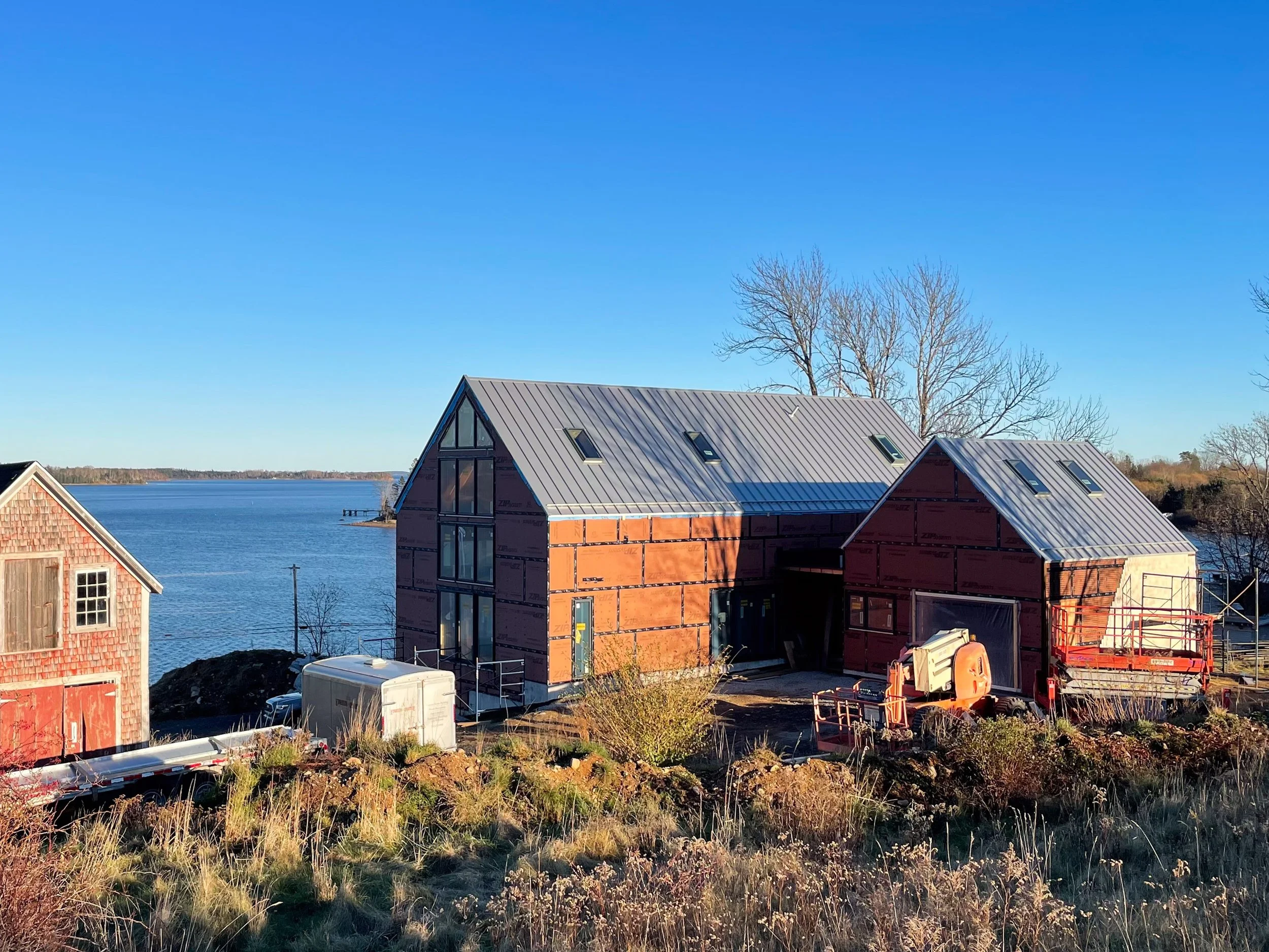 Under construction buildings near a body of water with a clear blue sky and leafless trees in the background.