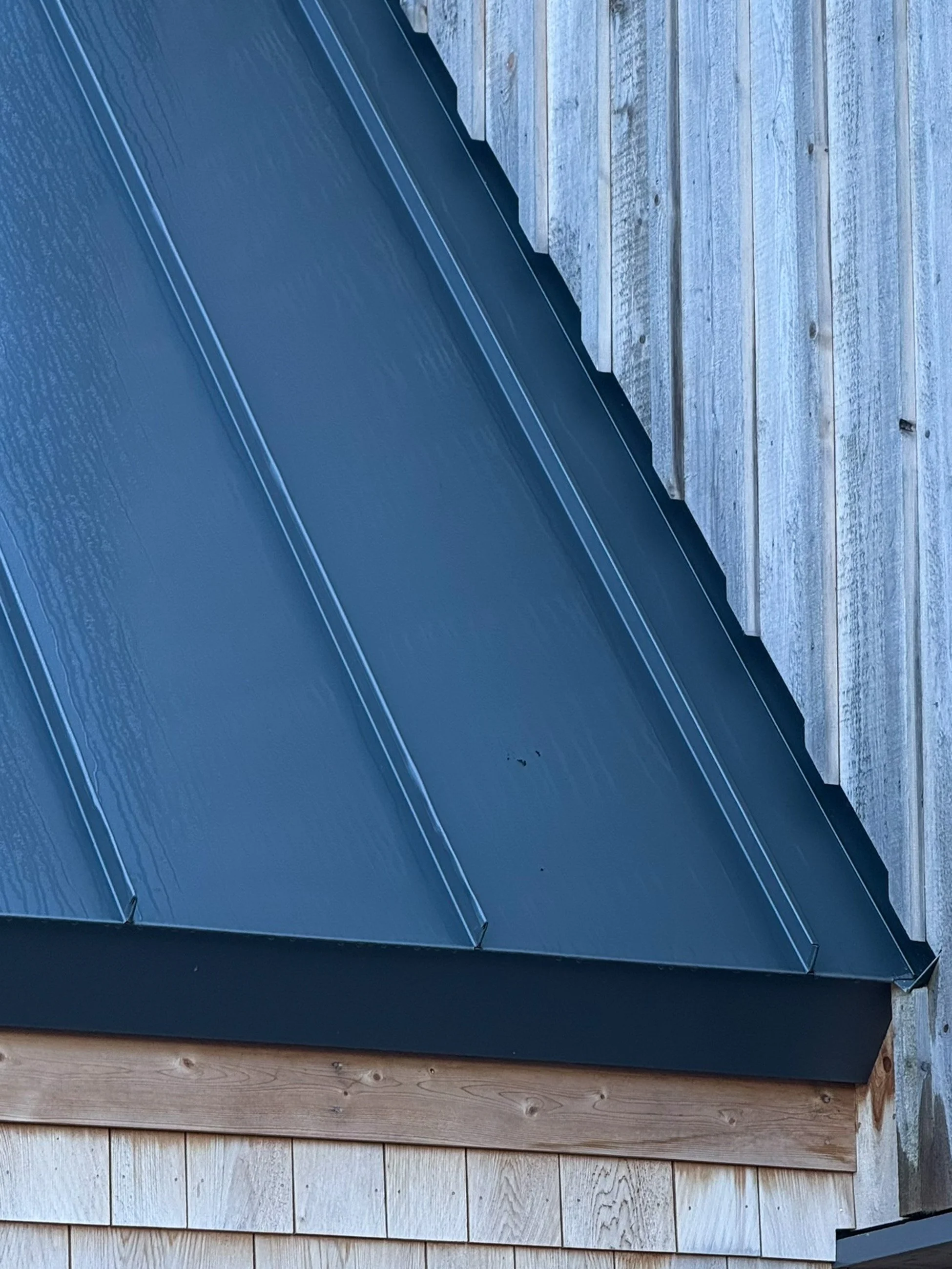 Close-up of a black metal roof with standing seam panels, adjacent to weathered wooden siding of a building.