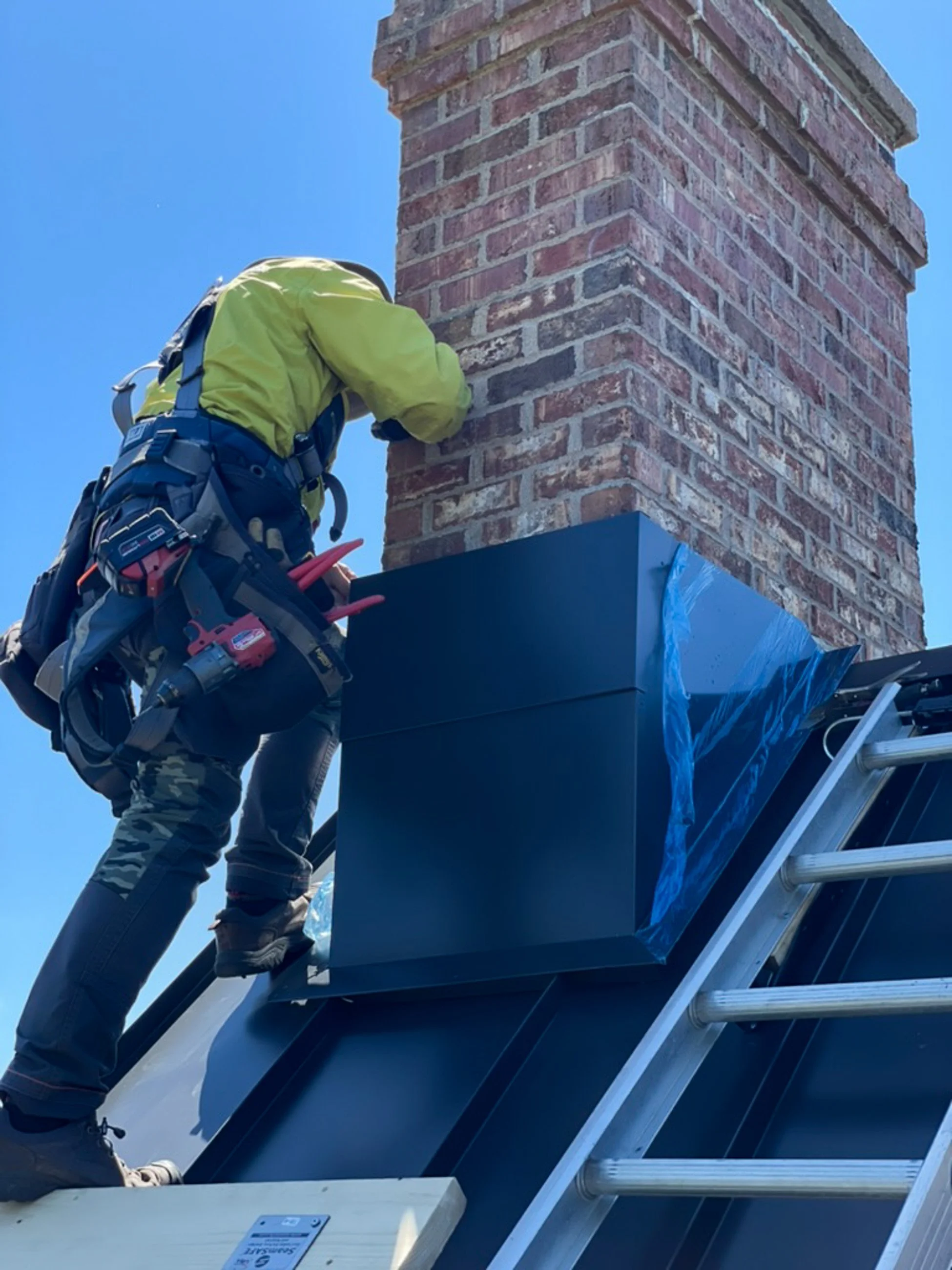 A worker installing a black chimney cap on a brick chimney, standing on a ladder, with tools attached to their belt, against a clear blue sky.