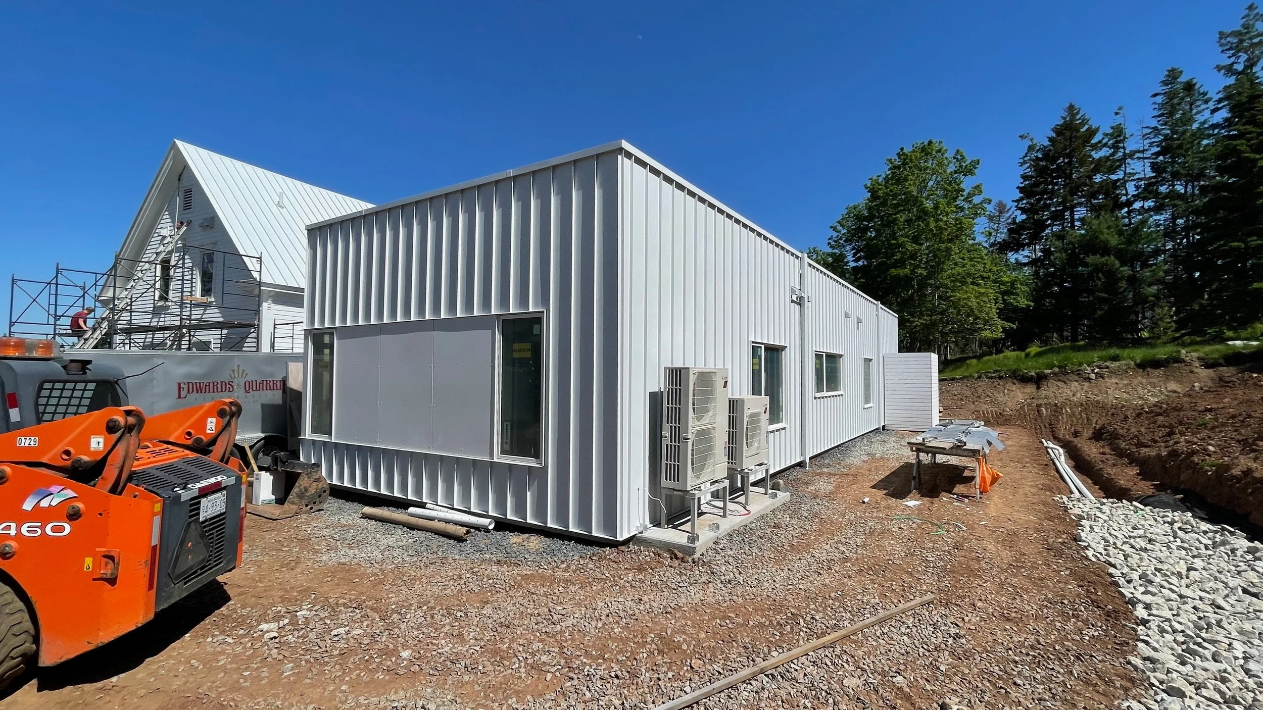 Construction site with a white metal building and equipment, with a second building under construction in the background, surrounded by trees and blue sky.