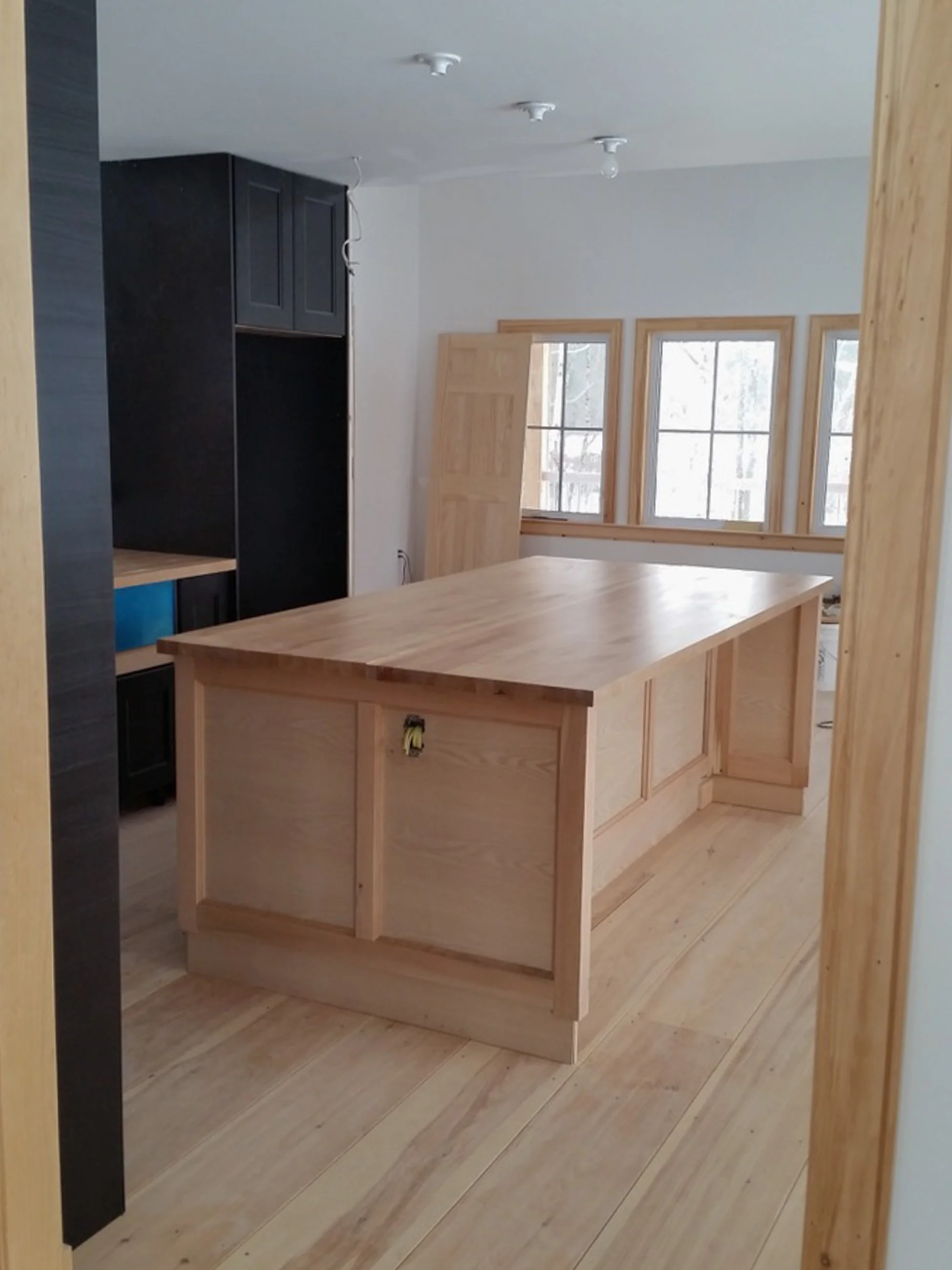 Interior of a kitchen under construction with a wooden island, three windows, and black cabinetry.