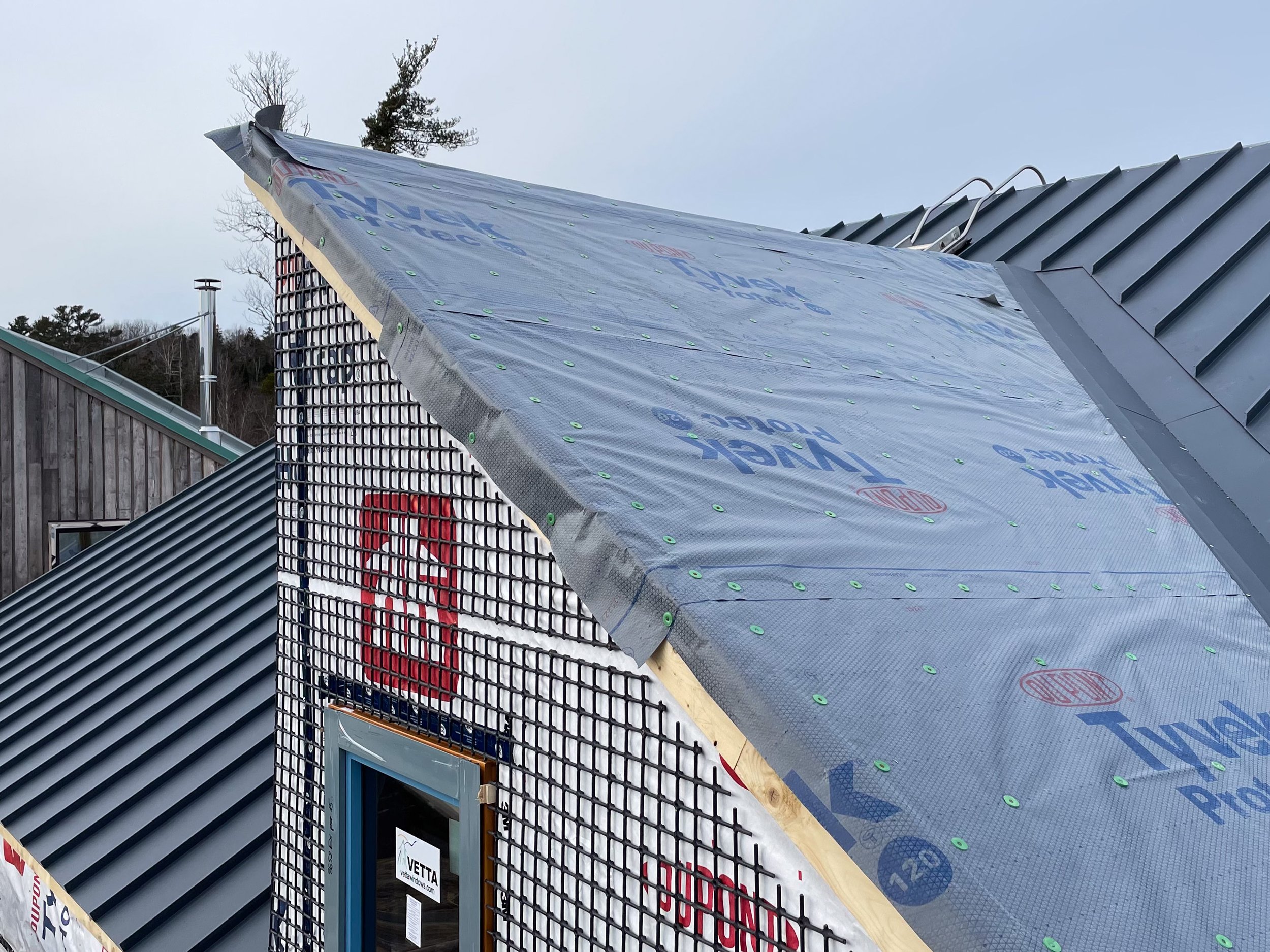 A house under construction with a partially installed metal roof. The exterior wall shows an underlayment material labeled 'Tyvek', and a skylight window is visible. The background includes trees and neighboring buildings.