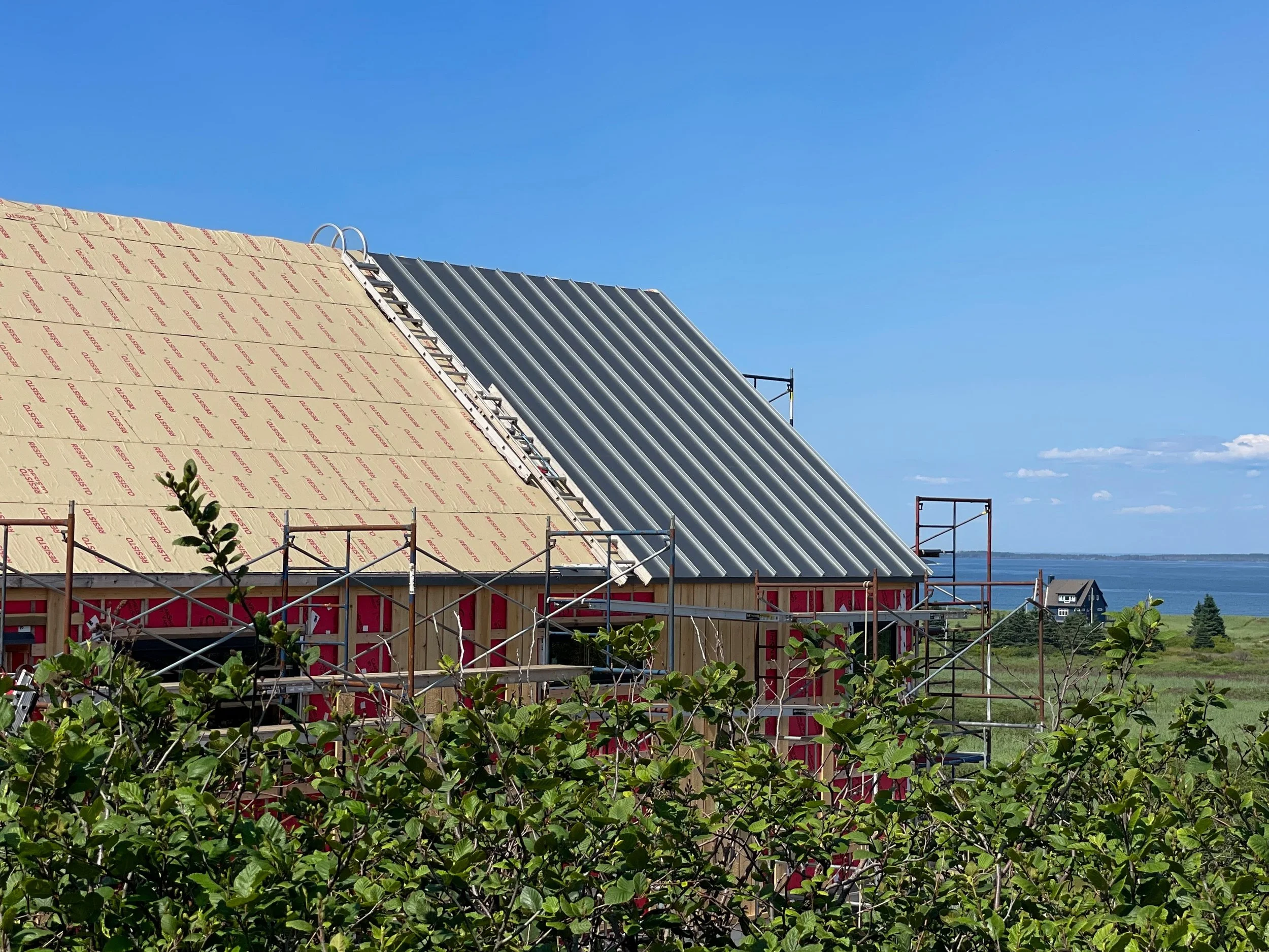 House under construction with metal roof, scaffolding, and blue sky, overlooking water and distant houses.