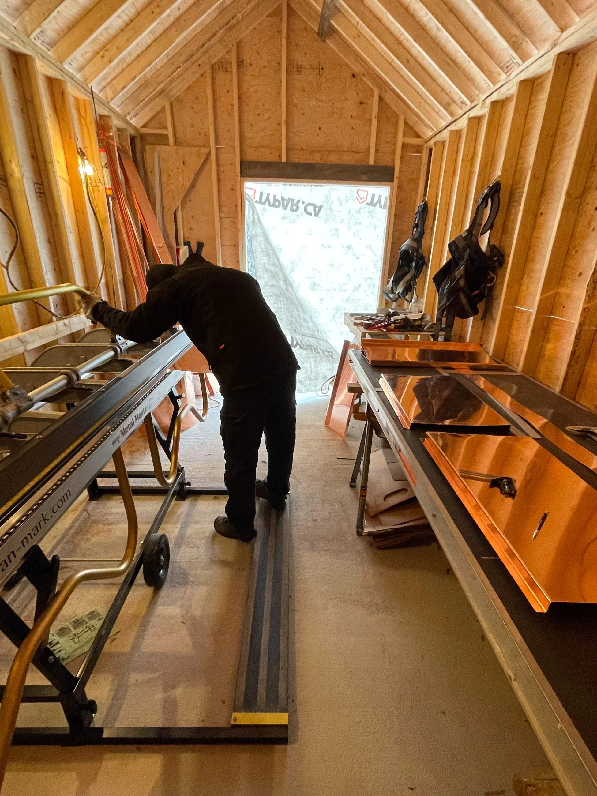 A person working inside a wood-framed room with walls and ceiling unfinished. The person is leaning over a metal worktable, possibly cutting or measuring. Various tools and equipment are nearby, and there is a large window at the end of the room covered with plastic sheeting.