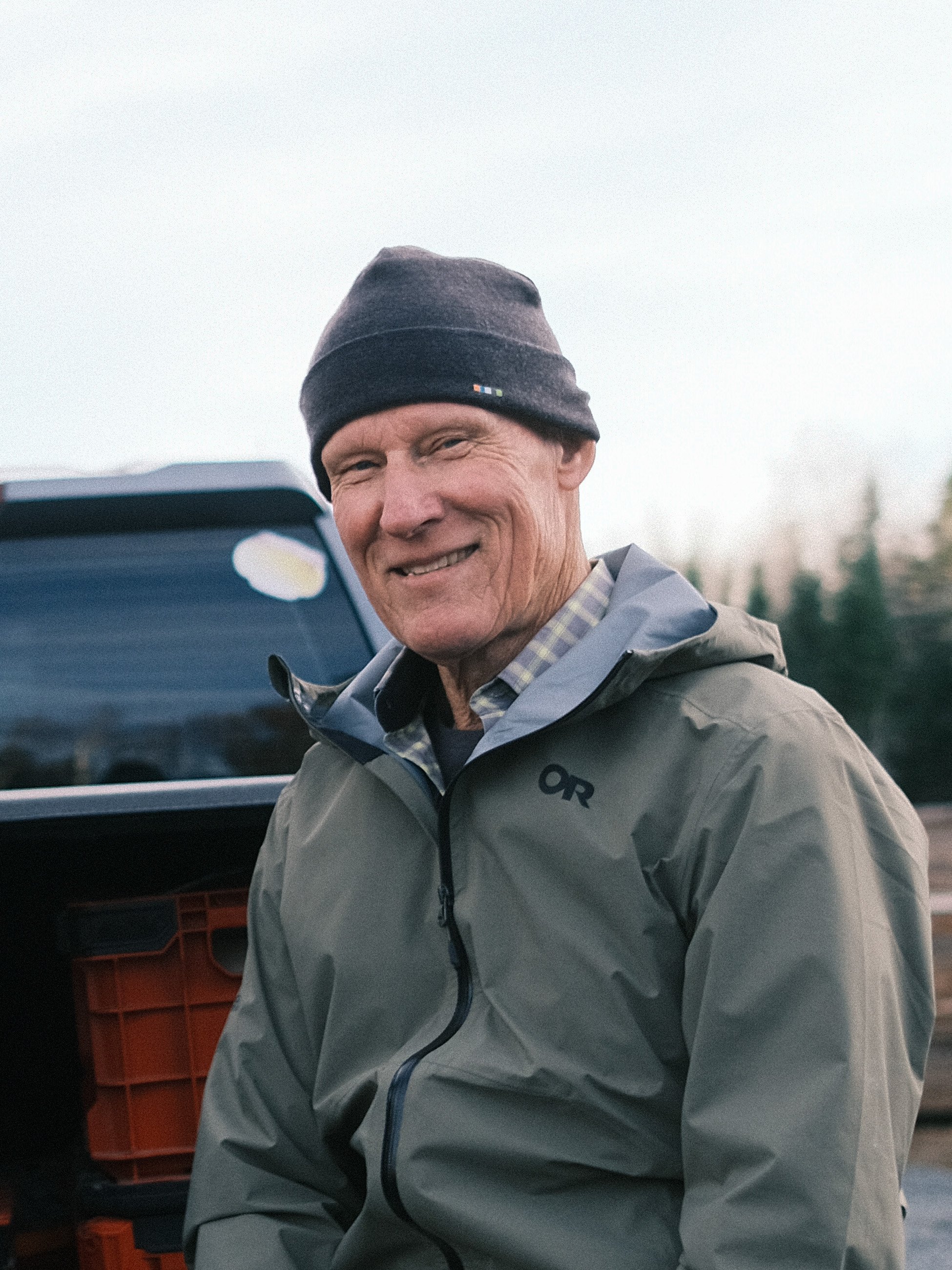 Man in a gray outdoor jacket and black beanie smiling outdoors near a vehicle with a background of trees and an overcast sky.