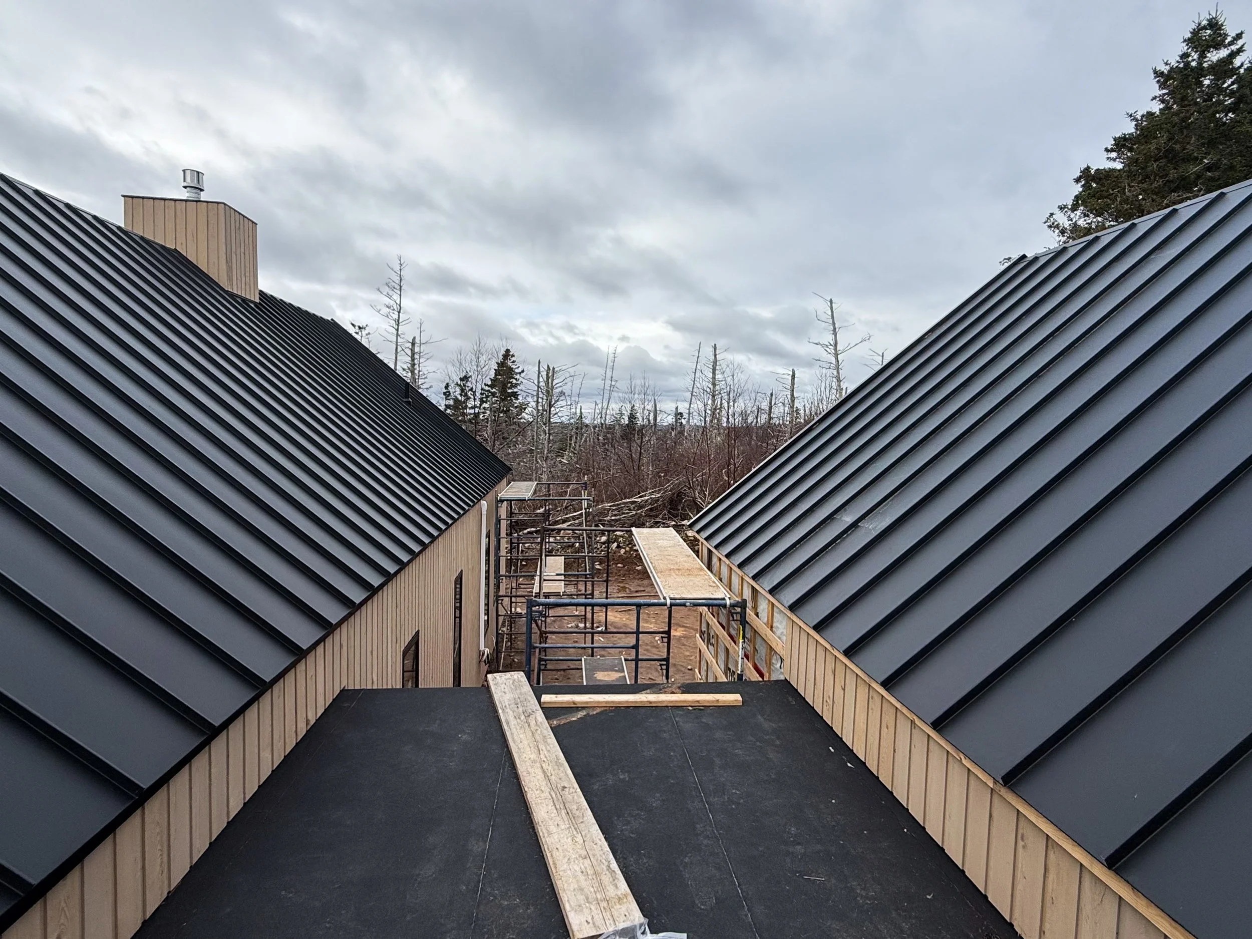 View of rooftops with black metal roofing, construction scaffolding, and wooden framing on a building under construction with overcast sky and trees in the background.