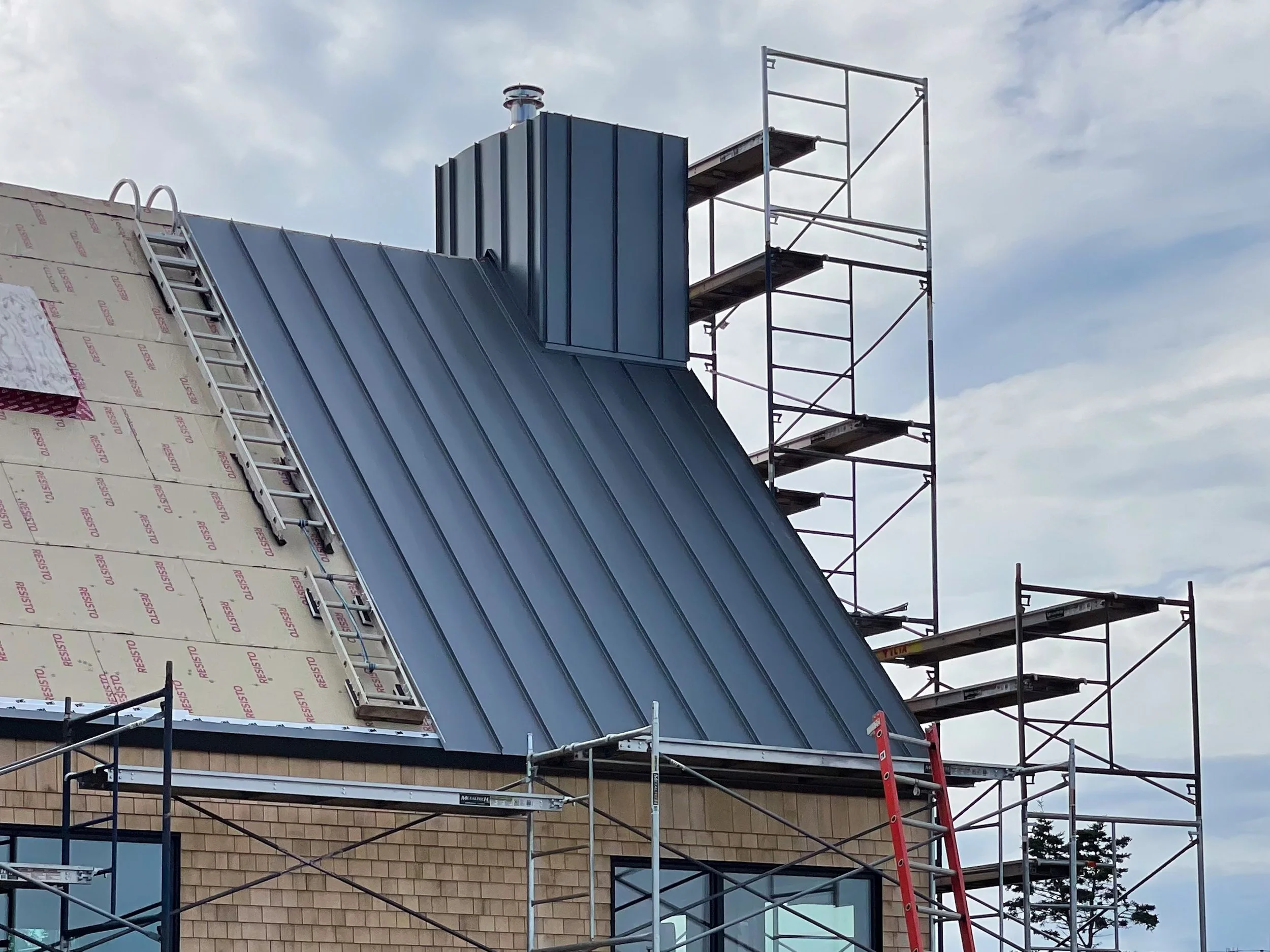 Construction scaffolding around a building with a new metal roof being installed. The sky is partly cloudy.