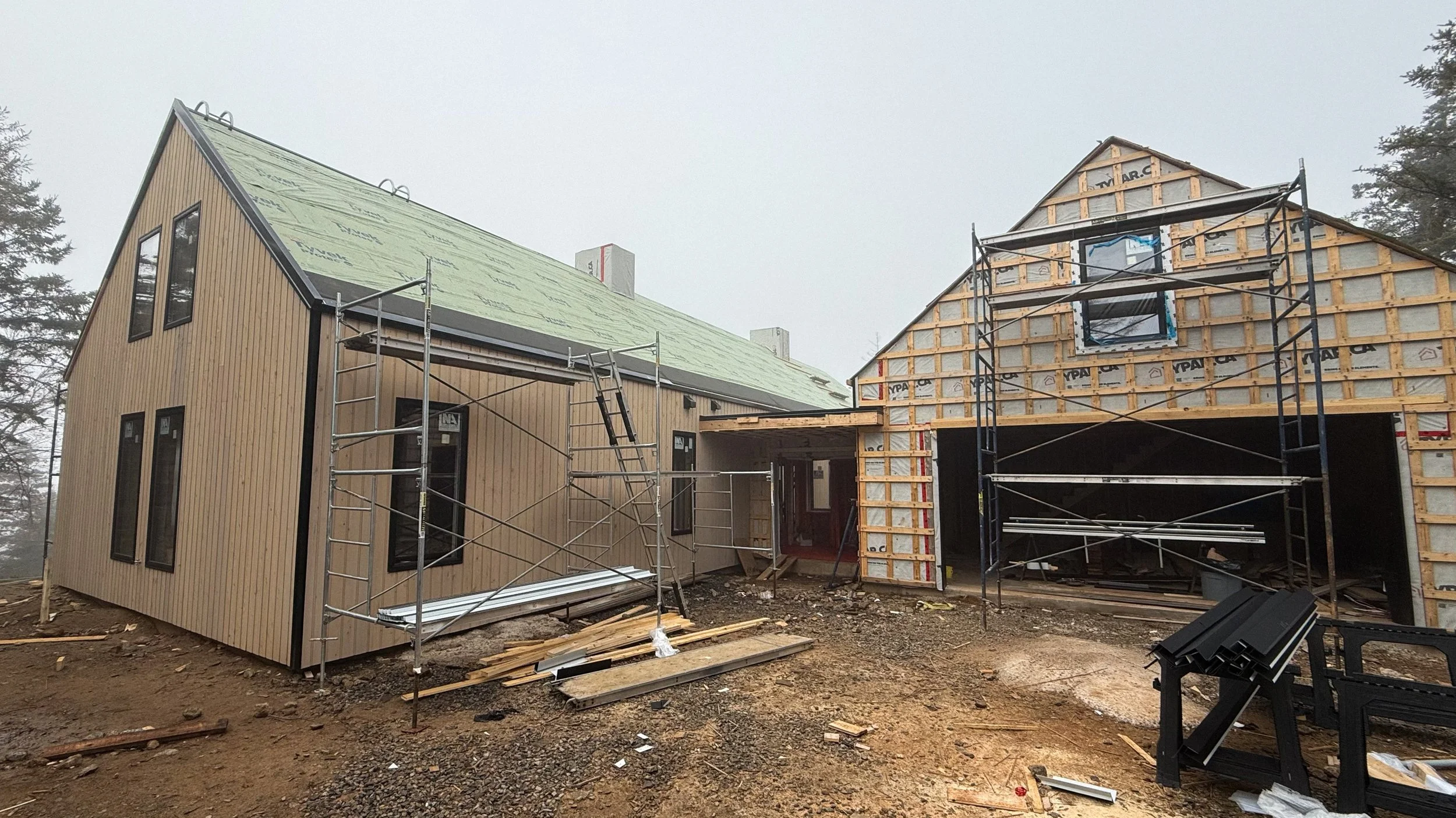 Two houses under construction, with scaffolding around them. The house on the left has exterior siding partially installed, and the house on the right is in the framing stage. Construction materials and tools are scattered on the ground.