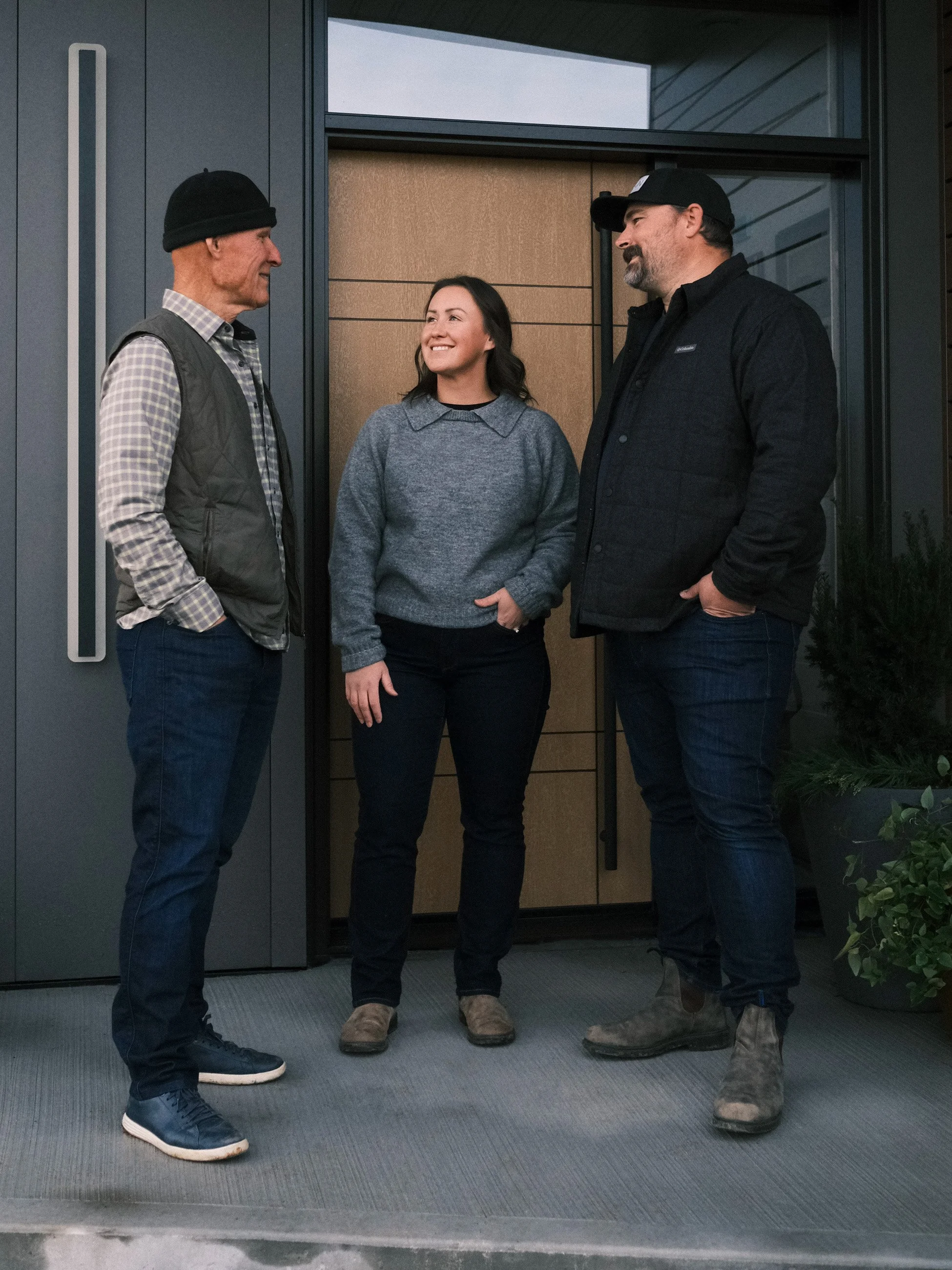 Three people standing and talking in front of a modern building entrance. The group includes two men and one woman, dressed casually, smiling and engaged in conversation.