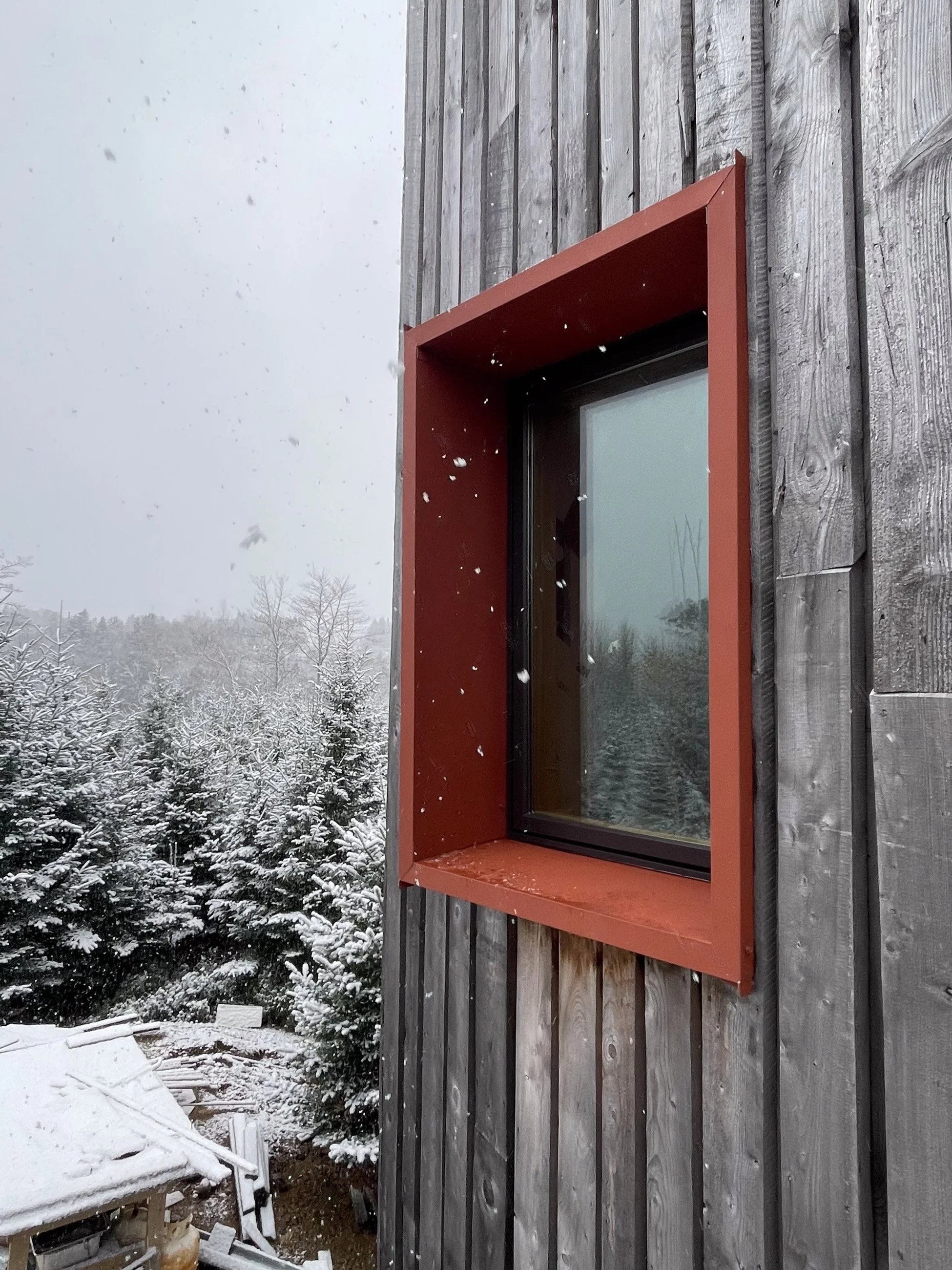 Close-up of a modern window with a reddish-brown frame on a weathered wooden wall, snow falling in a winter landscape with snow-covered trees in the background.