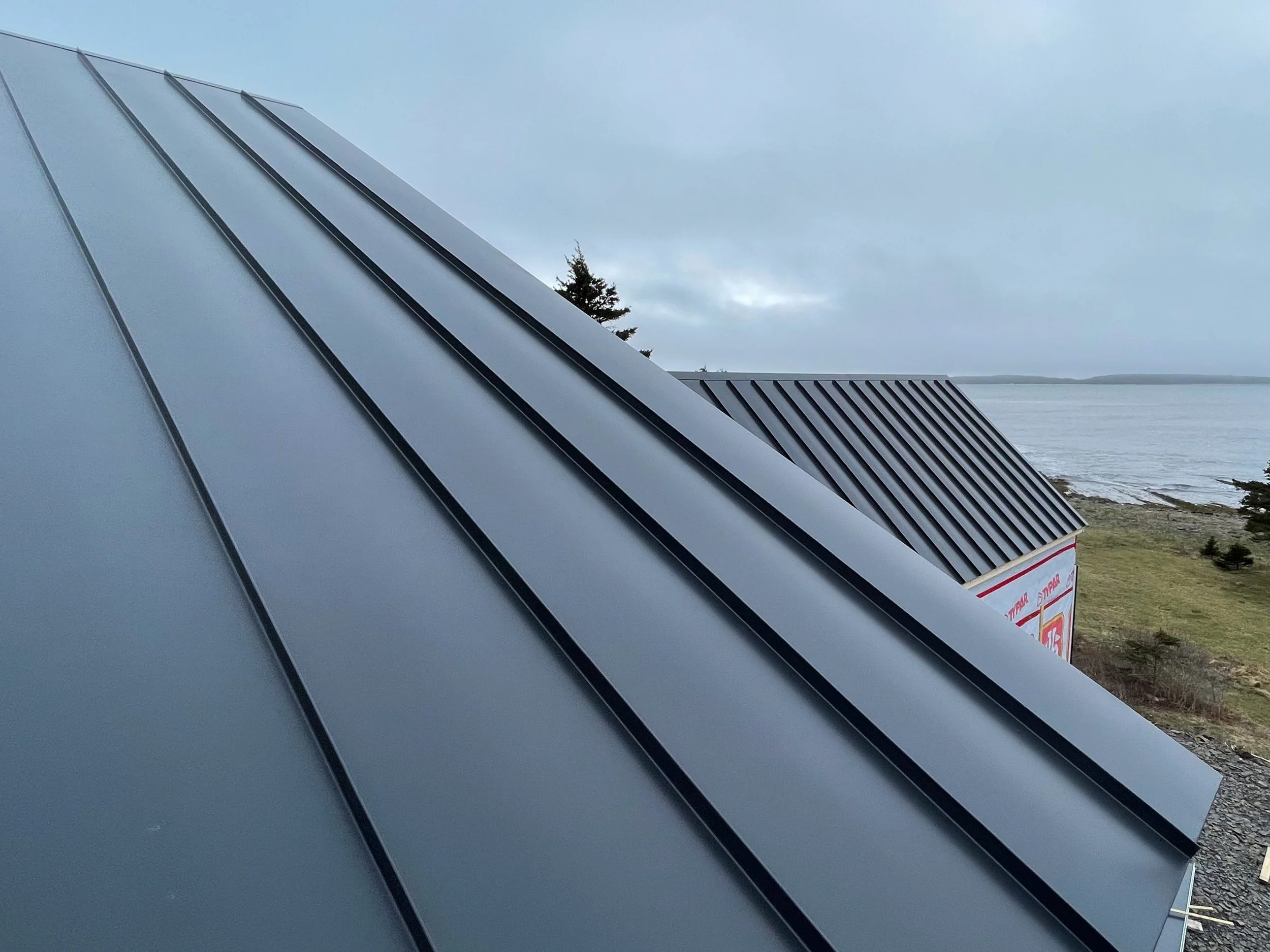Close-up of blue metal roofing panels on a building with a view of a body of water and a cloudy sky in the background.
