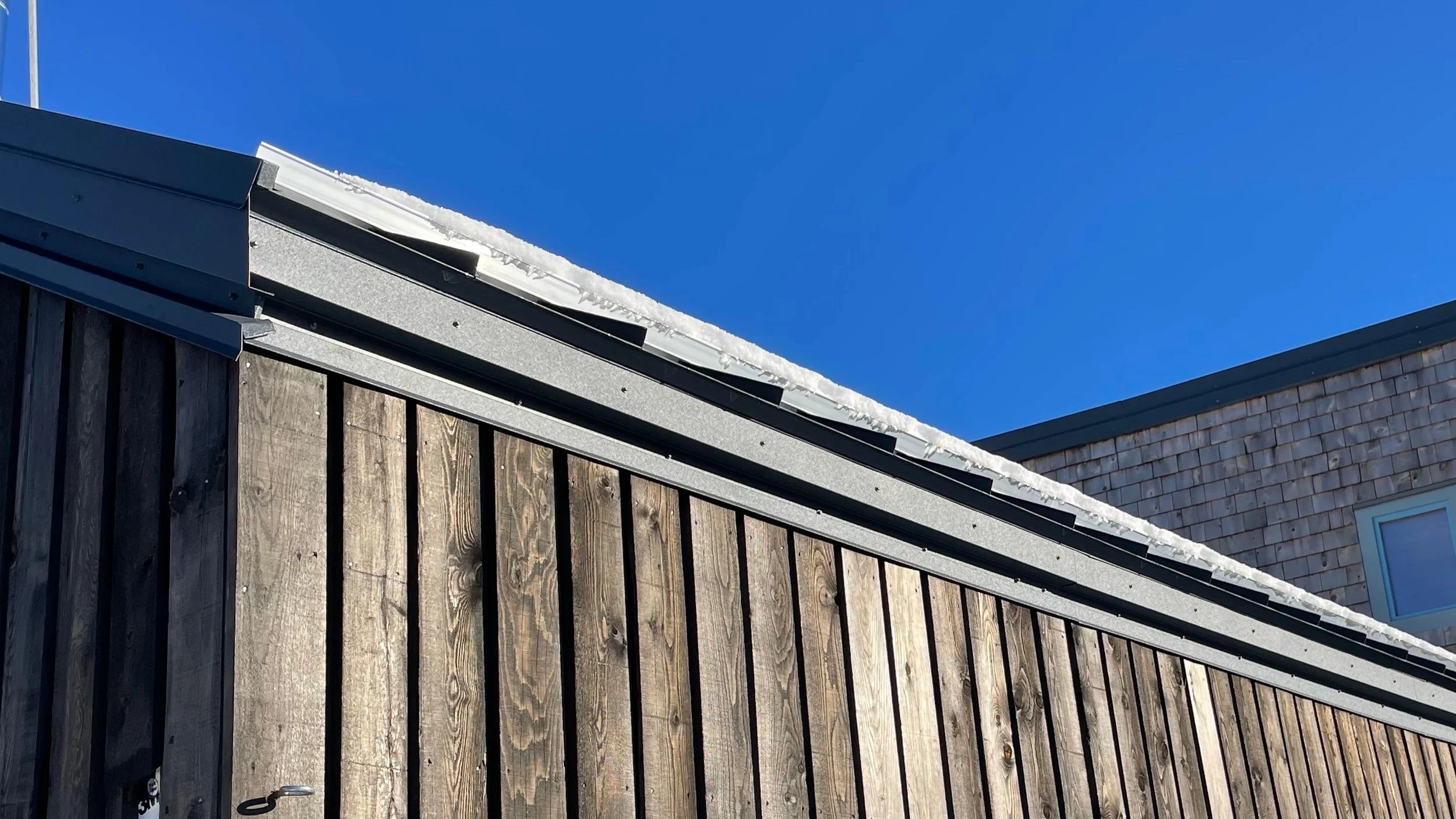 A rooftop with snow and ice along the edge, wooden exterior walls, a metal gutter, and a house with shingles and a window in the background against a clear blue sky.
