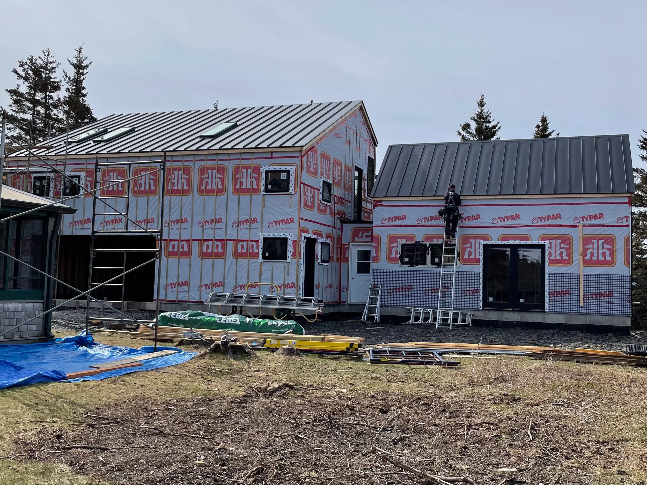 Construction site with two houses under construction, scaffolding, ladders, and construction worker, surrounded by building materials and equipment.