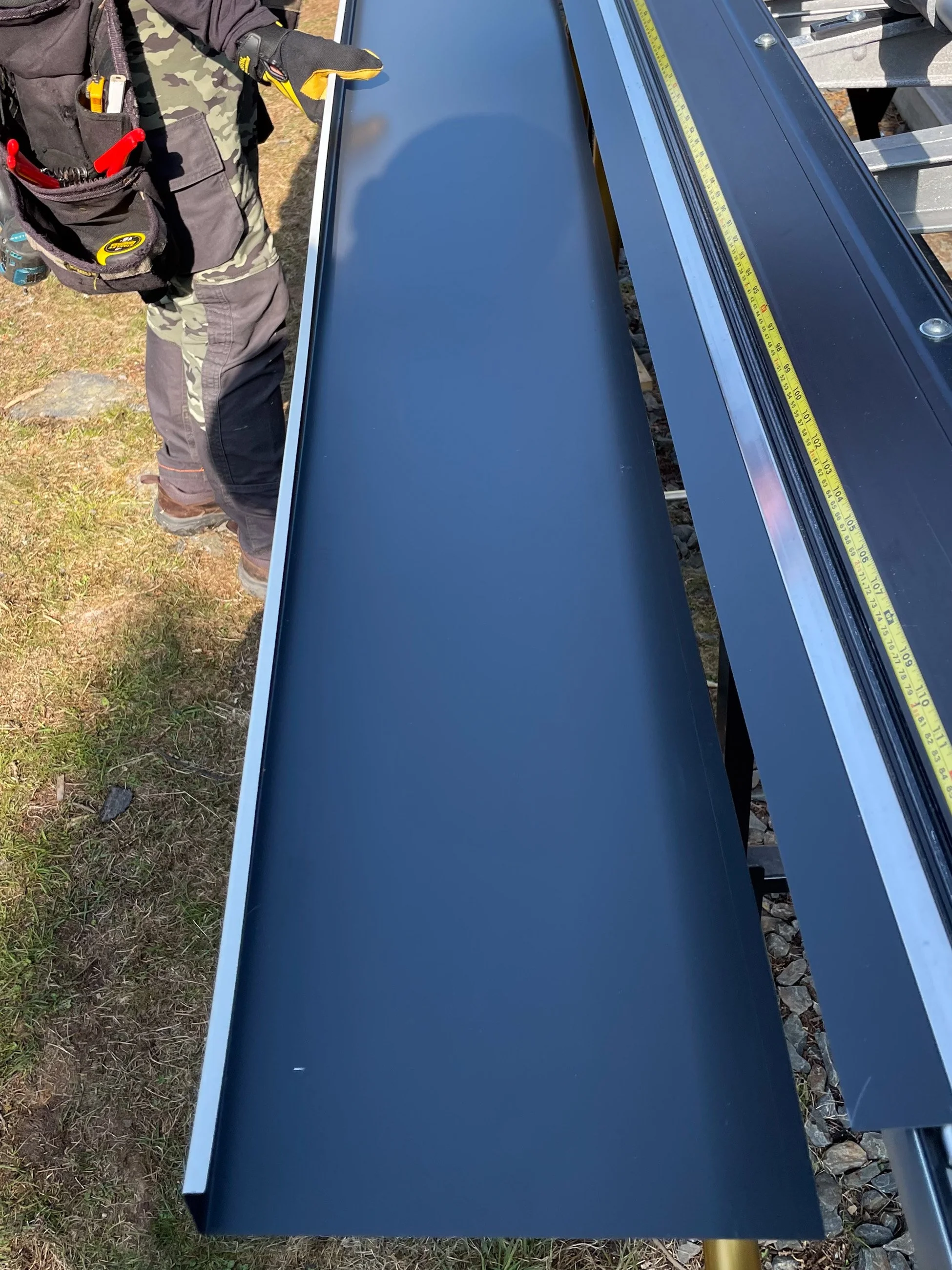 Close-up of a metal work surface or table next to a person wearing work gloves and a tool belt, set outdoors on grass, with a yellow measuring tape on the surface.
